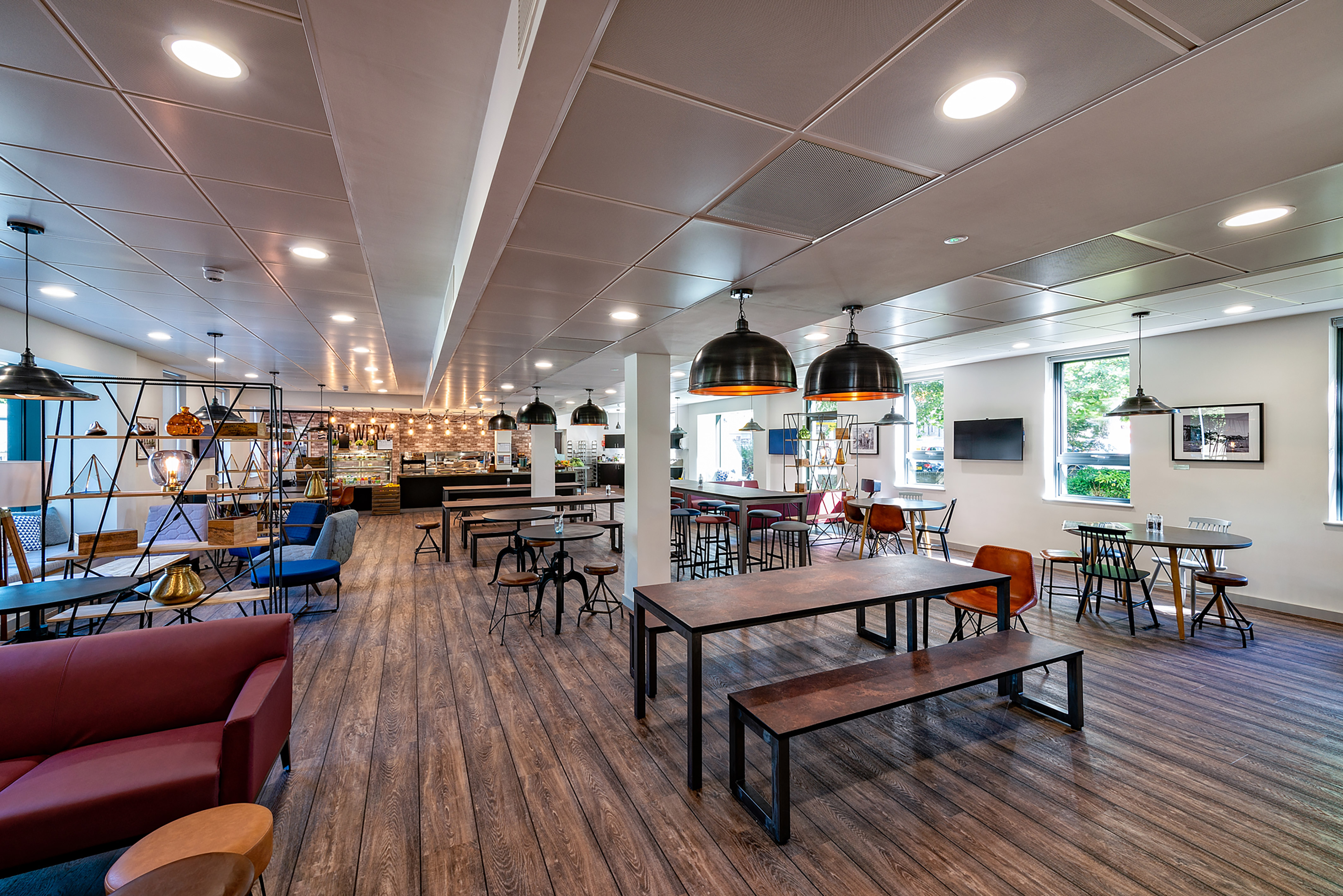 Spacious and modern cafeteria or communal dining area featuring wooden floors, various seating options, pendant lighting, and a well-lit open layout