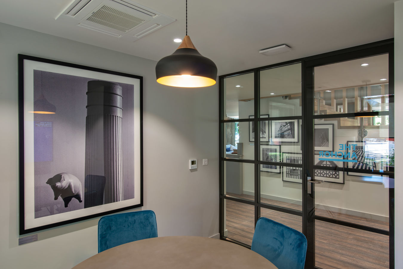 Framed artwork of a column and pig decorates a modern office room; a hanging pendant light illuminates blue chairs and a round table, with glass partitions displaying "THE ANCHOR" text.