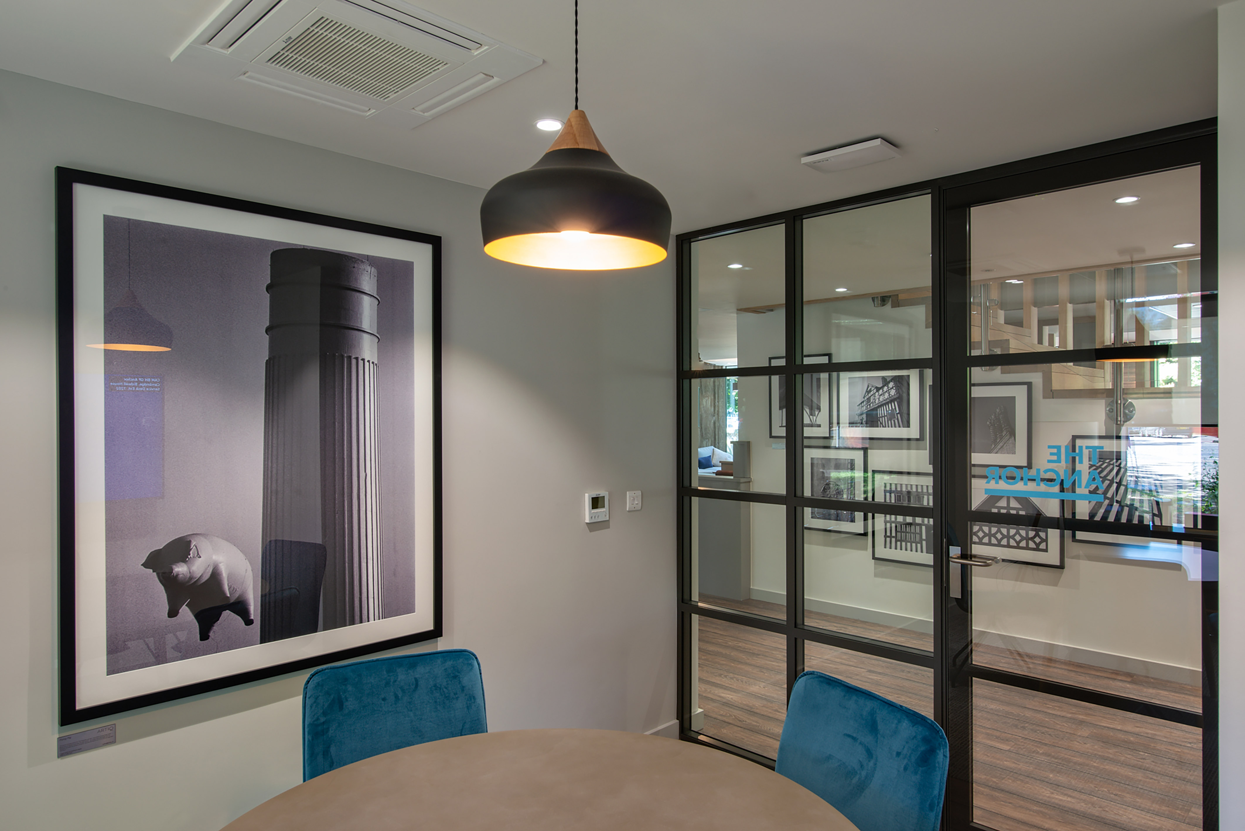 Framed artwork of a column and pig decorates a modern office room; a hanging pendant light illuminates blue chairs and a round table, with glass partitions displaying "THE ANCHOR" text.