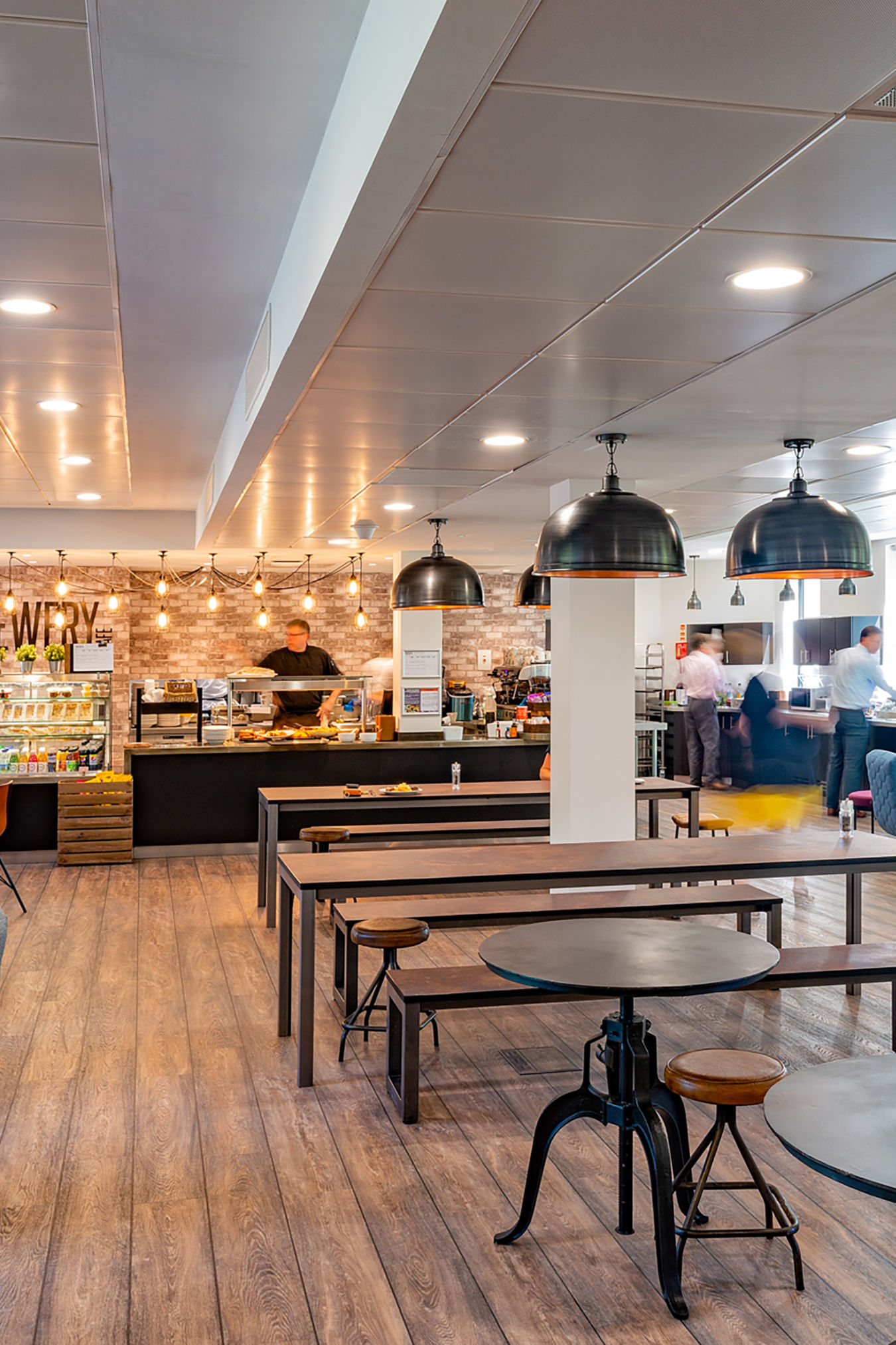 Spacious, modern cafe with wood flooring, industrial lighting, and varied seating. Cozy and inviting ambiance, with a bustling counter in the background