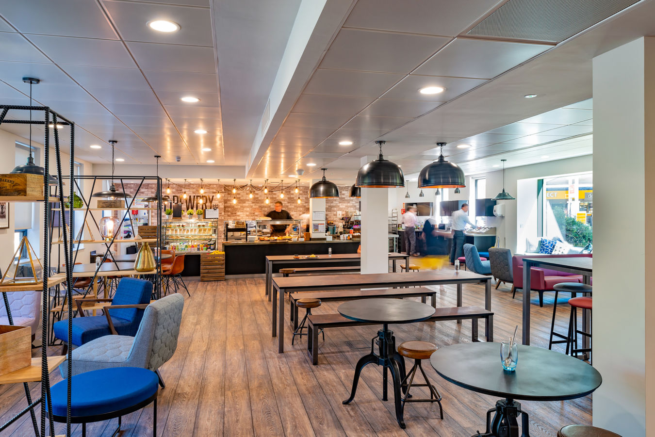 Spacious, modern cafe with wood flooring, industrial lighting, and varied seating. Cozy and inviting ambiance, with a bustling counter in the background