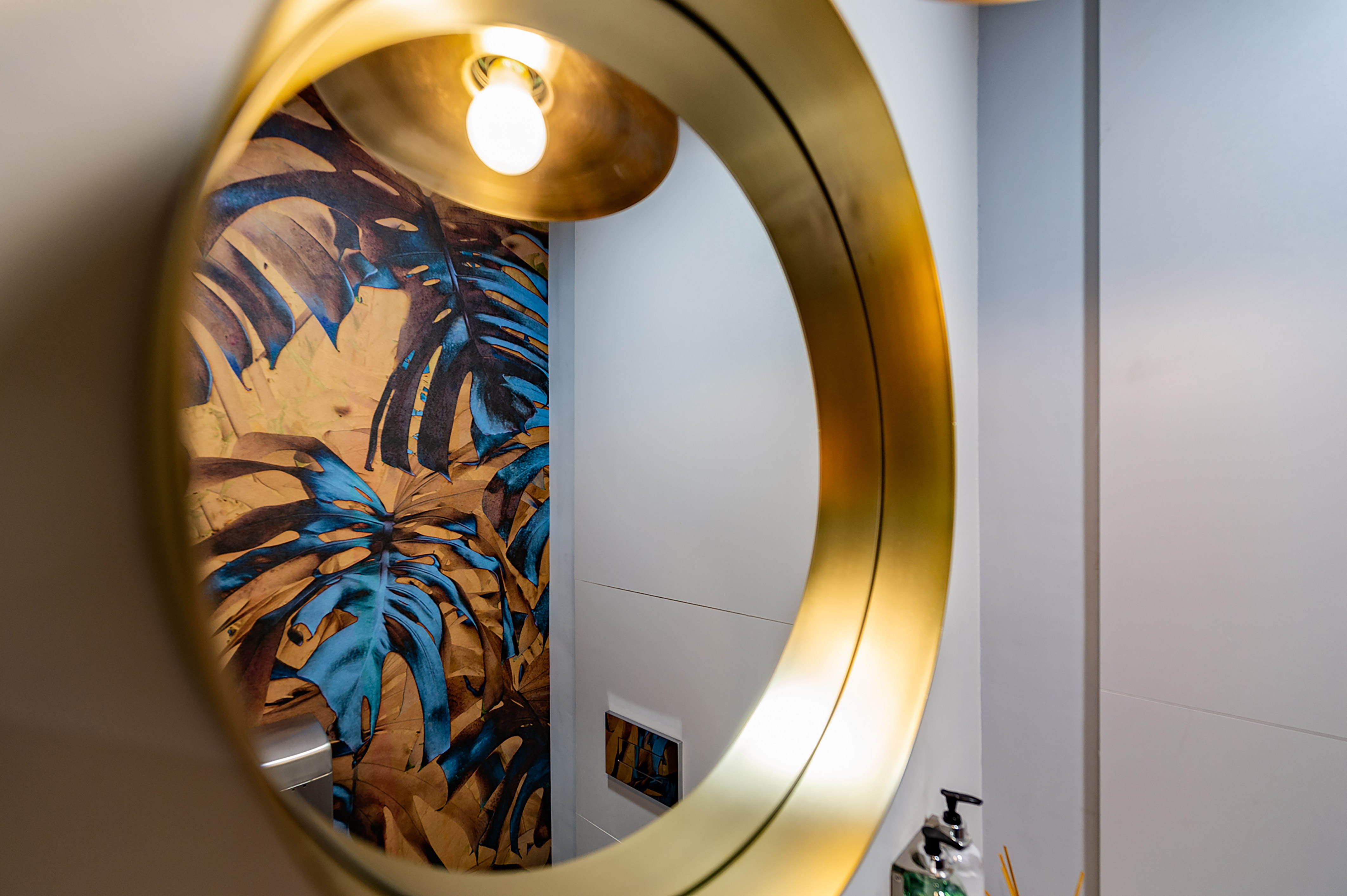 A round mirror with a golden frame reflects a vibrant wall with blue and brown tropical leaf patterns; a lit bulb hangs above and toiletries are nearby in a modern bathroom.