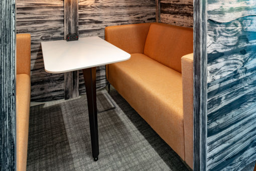 Compact office booth with an orange upholstered bench, white table, and wood-textured partition walls.