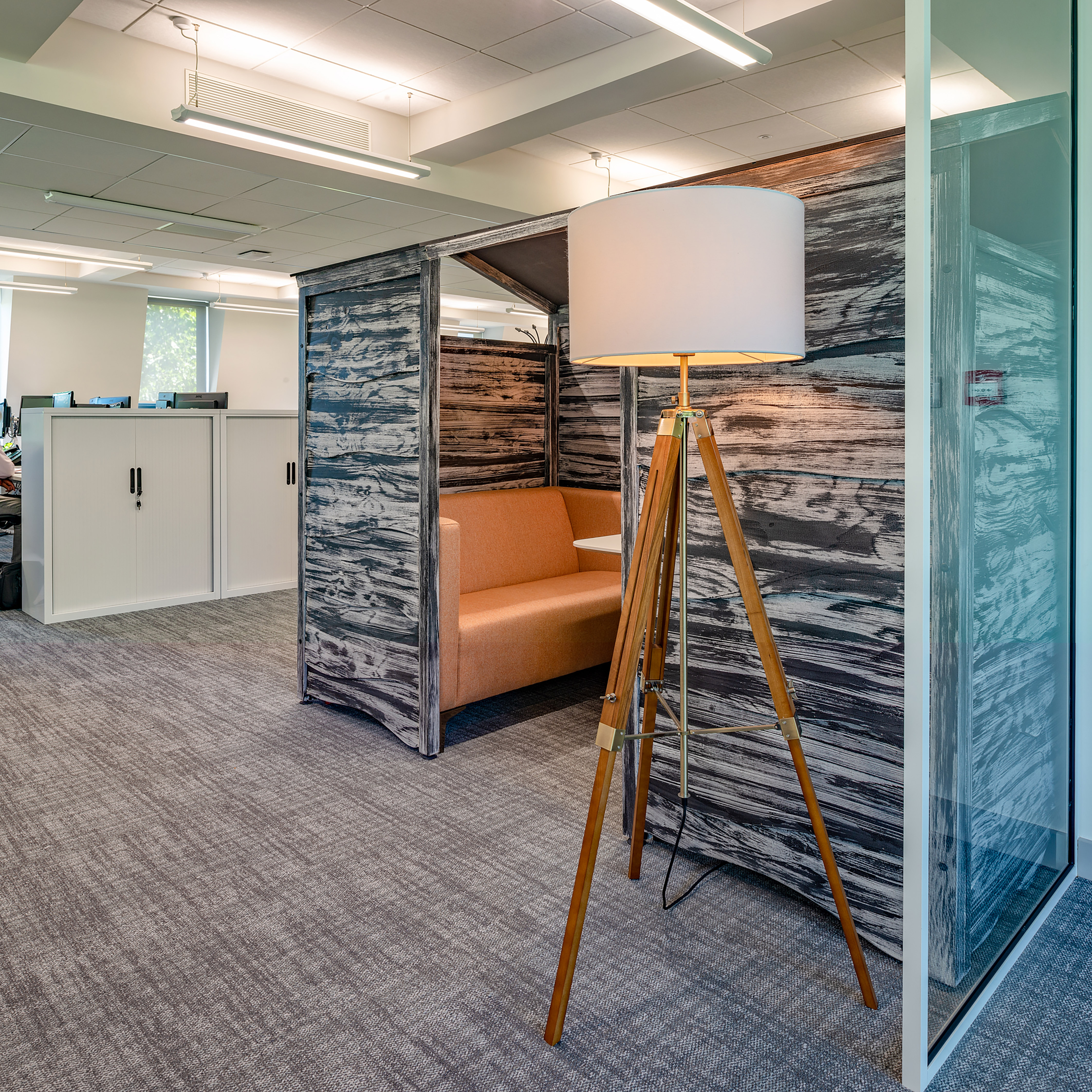 Modern office space with a cozy nook featuring a rustic wood-patterned wall and an orange cushioned bench. A tripod floor lamp adds warmth