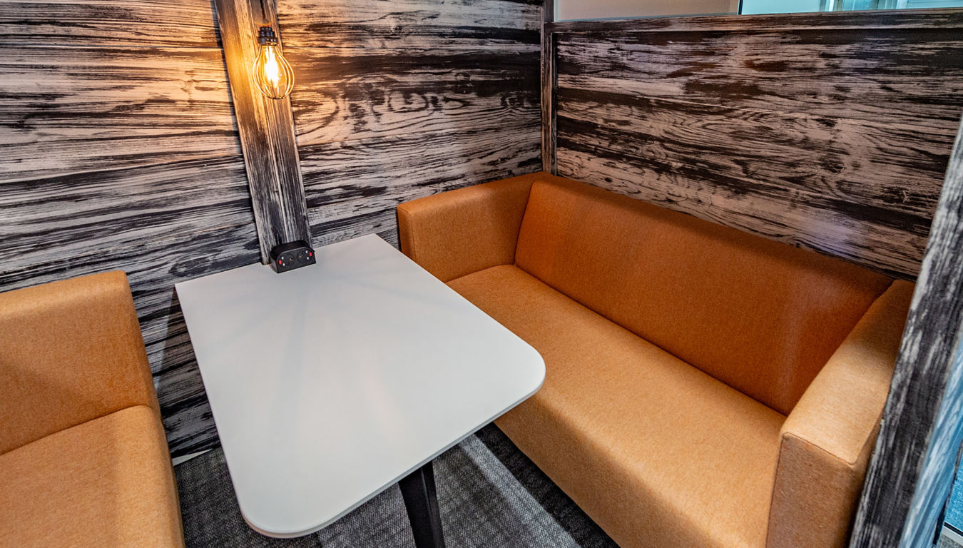 Cozy booth with a white table, tan cushioned seating, and a warm exposed bulb. Wood-paneled walls create a rustic, inviting atmosphere