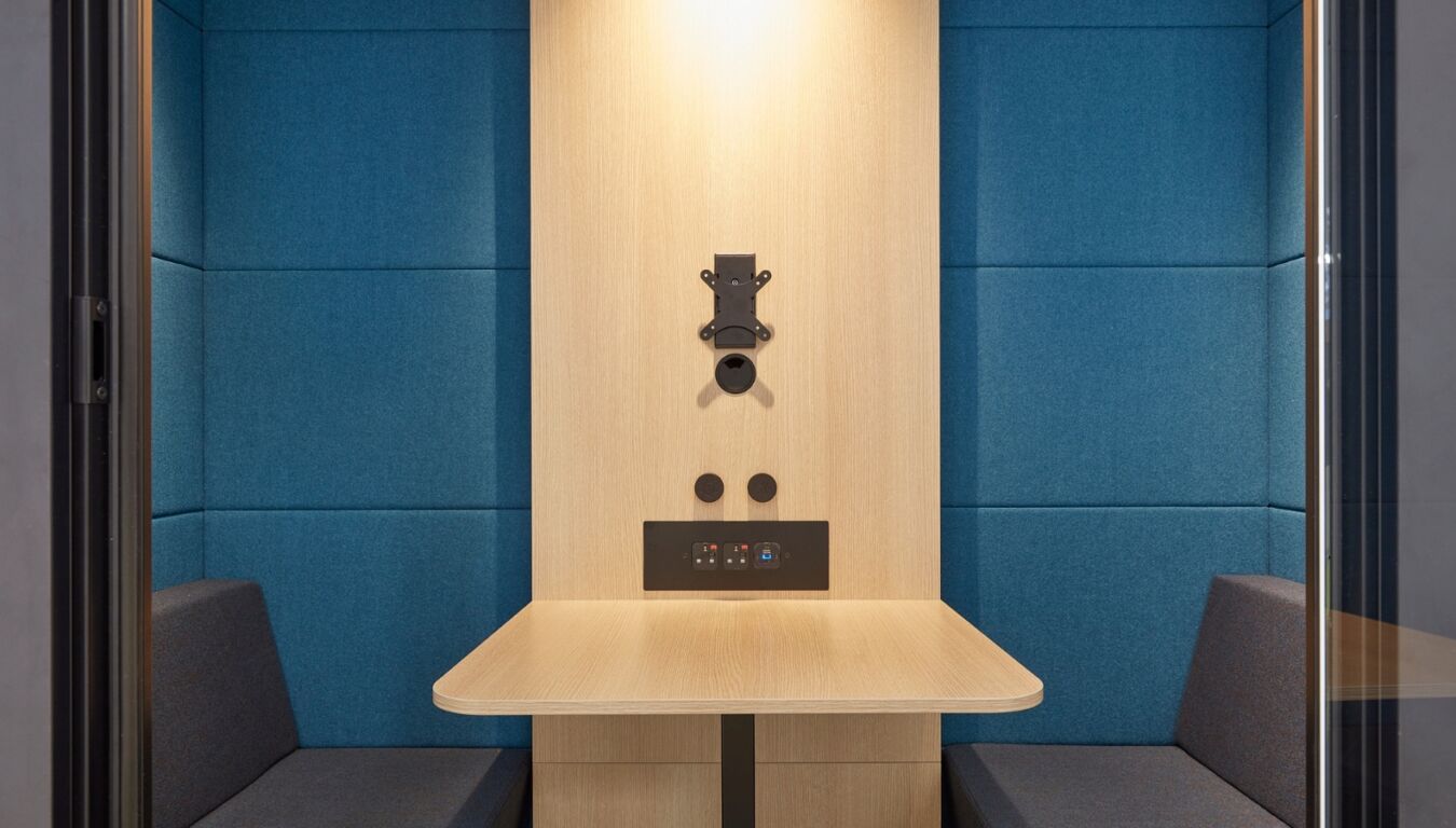 A modern, minimalistic booth features a wooden table, wall-mounted equipment, and two cushioned benches. The blue upholstered walls create a cozy, enclosed space ideal for individual work or meetings.