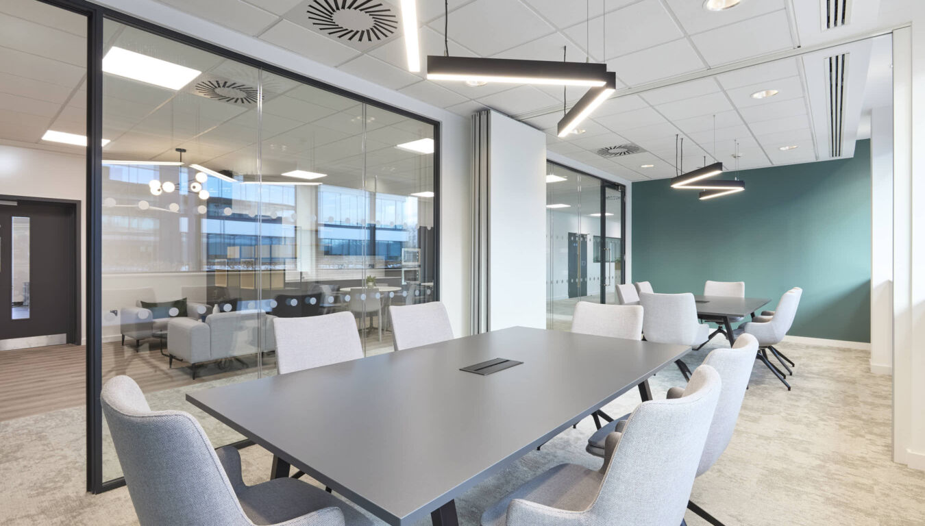 Modern conference room with gray table and chairs, illuminated by sleek pendant lights; glass walls reveal additional seating and a teal accent wall in a bright office setting.