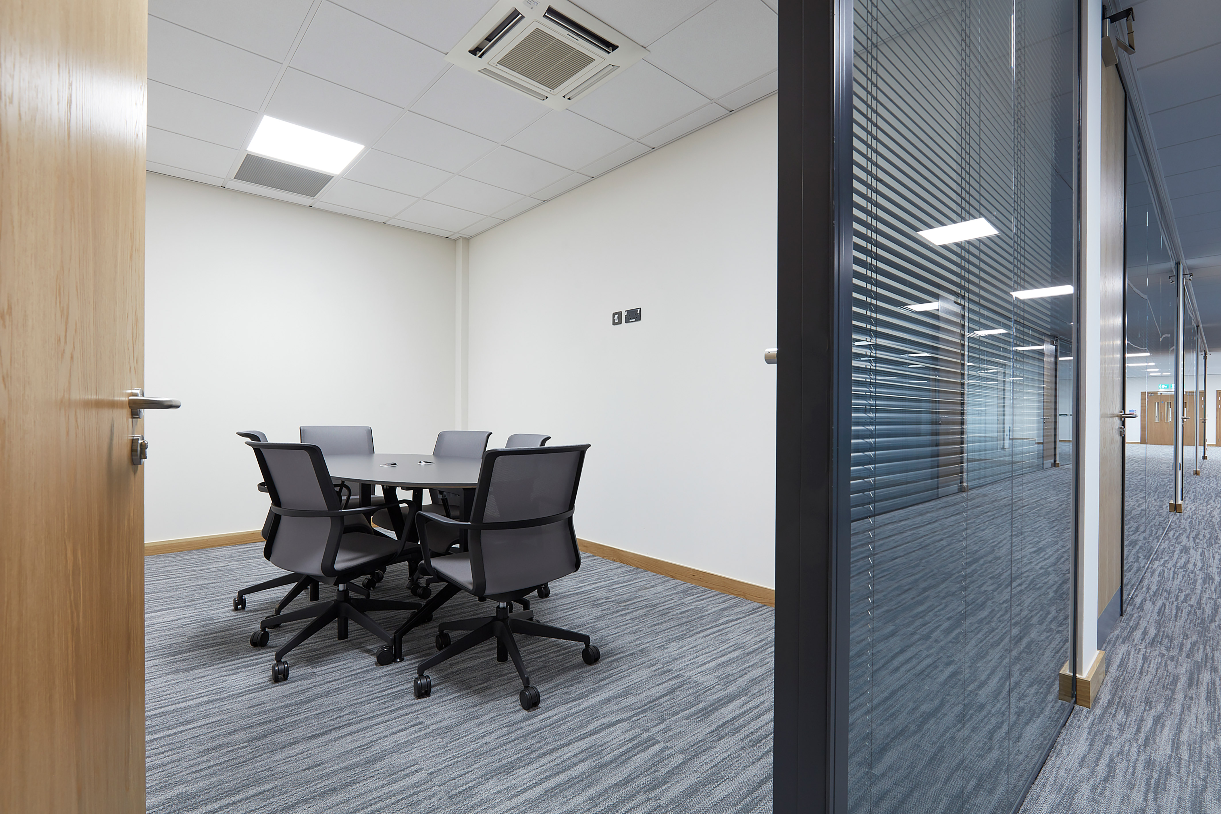 Small meeting room with a rectangular table and five mesh chairs on wheels. Glass wall with blinds and a ceiling light. Calm, professional tone