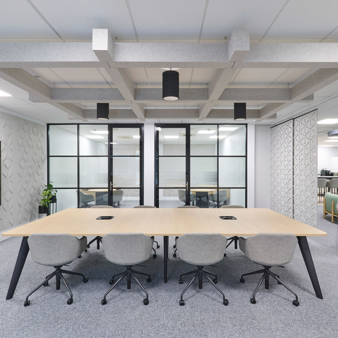 Minimalist conference room with a long wooden table, ergonomic chairs, dual wall-mounted screens, and glass partitions.