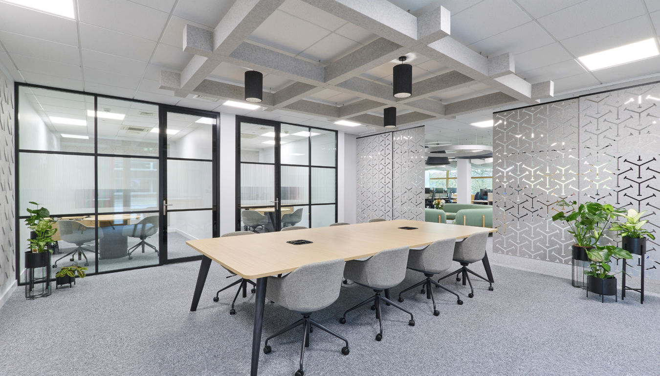 A conference table with eight grey chairs is arranged in a modern office, surrounded by glass partitions and geometric-patterned walls, with suspended ceiling decor and natural light through windows.