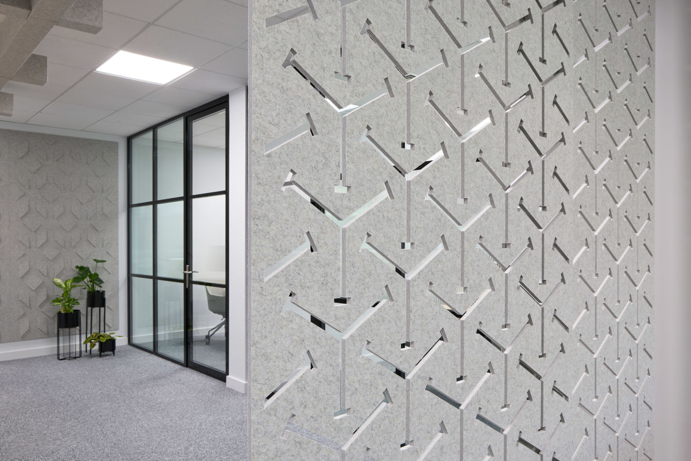 Decorative wall with geometric, arrow-shaped cutouts enhances an office space; glass partition door and indoor plants accentuate the modern, minimalist design.