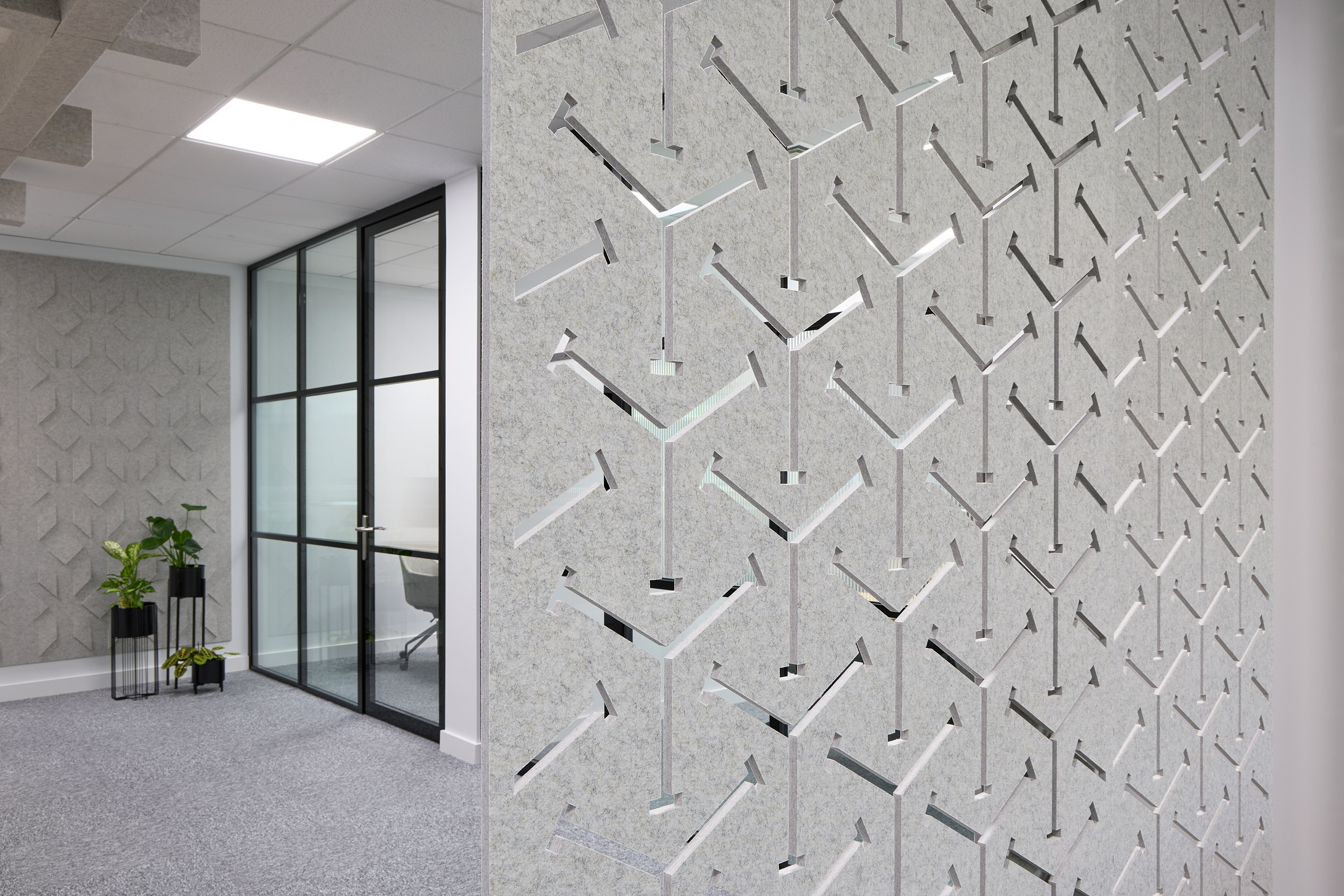 Decorative wall with geometric, arrow-shaped cutouts enhances an office space; glass partition door and indoor plants accentuate the modern, minimalist design.