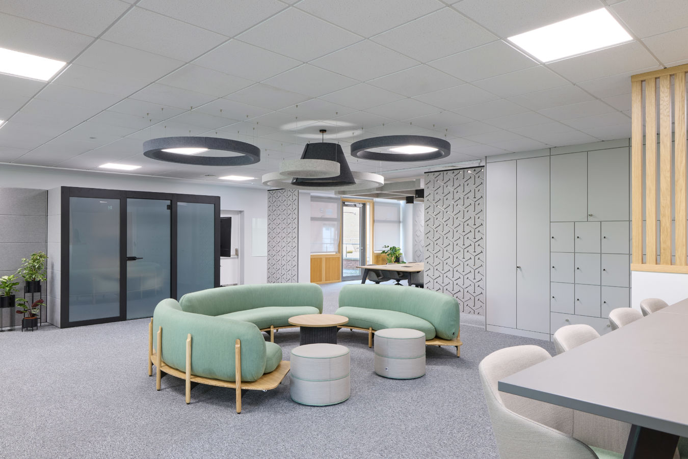 Modern office space featuring a curved green sofa set with round tables in the center, surrounded by chairs and lockers, under geometric ceiling lights on a gray carpeted floor.