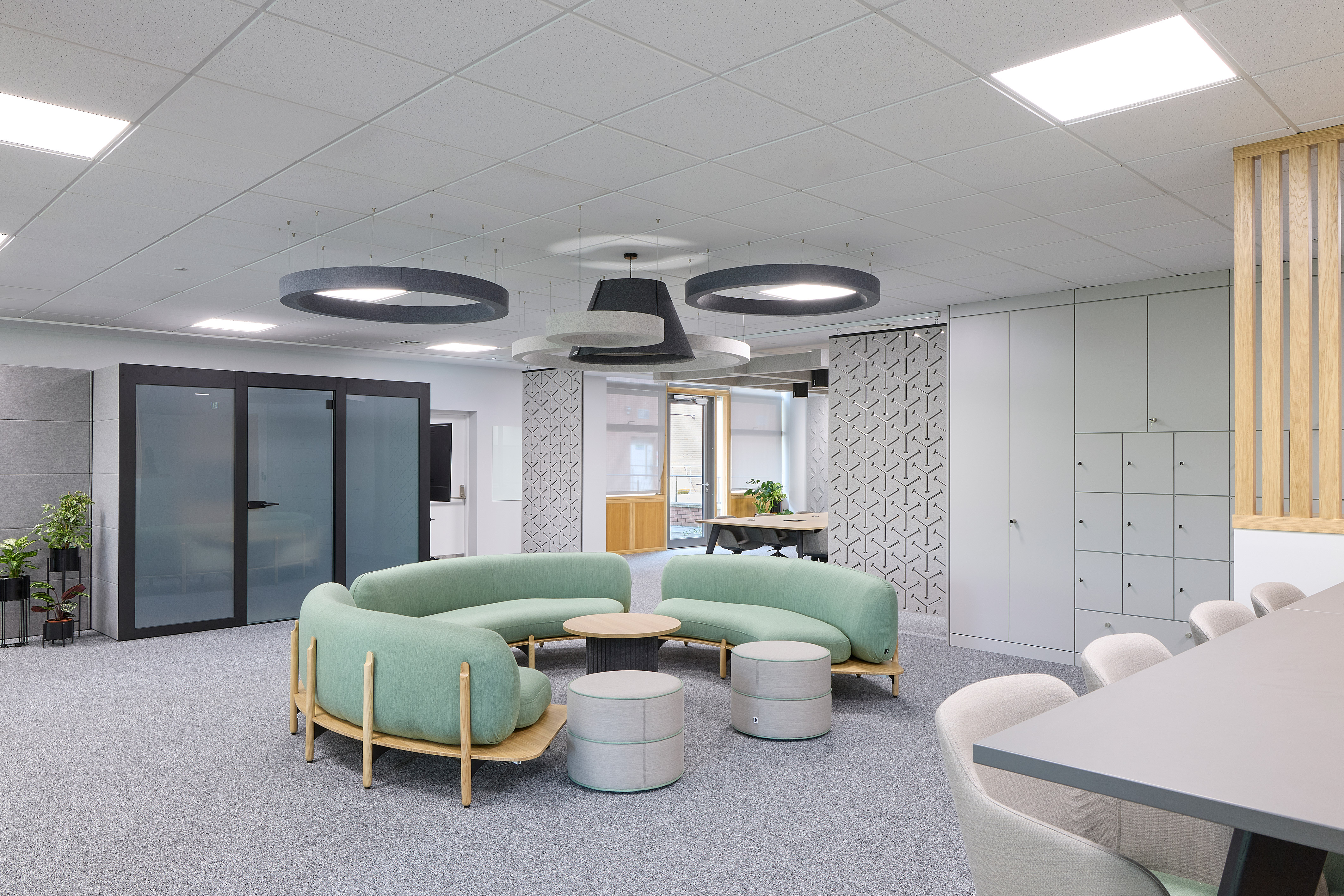 Modern office space featuring a curved green sofa set with round tables in the center, surrounded by chairs and lockers, under geometric ceiling lights on a gray carpeted floor.