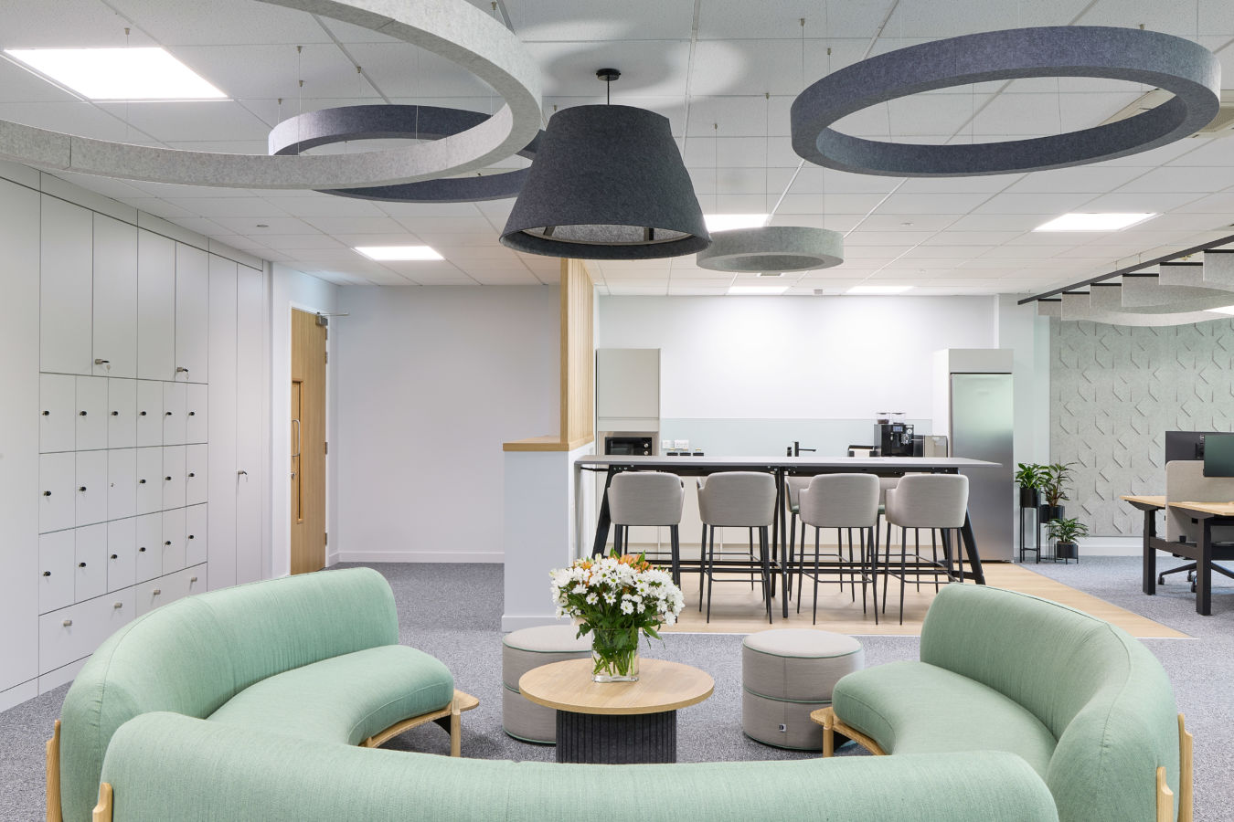 Curved mint-green sofas encircle a small round table with white flowers, set in a modern office space with lockers, high stools, and ceiling light fixtures.