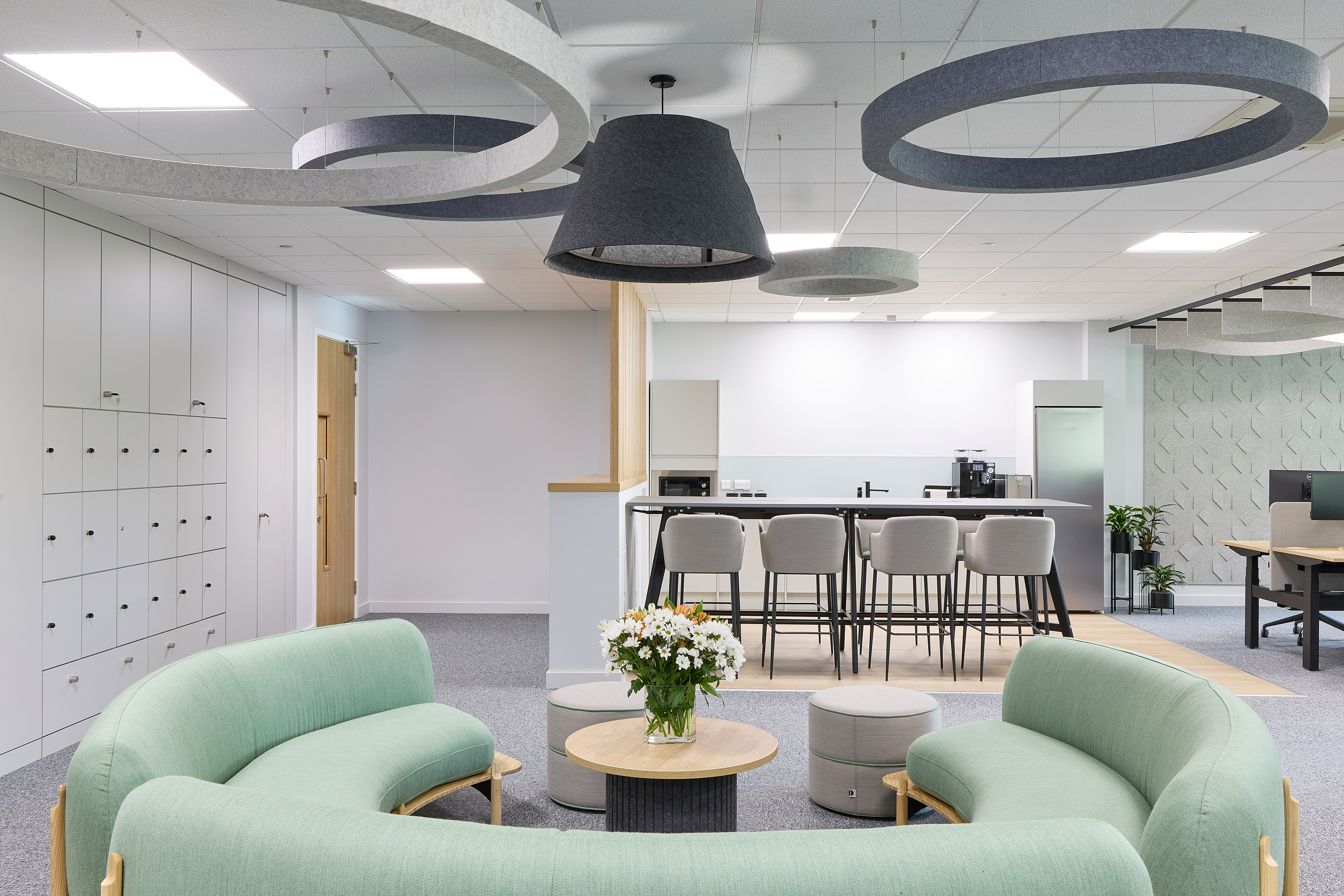 Curved mint-green sofas encircle a small round table with white flowers, set in a modern office space with lockers, high stools, and ceiling light fixtures.