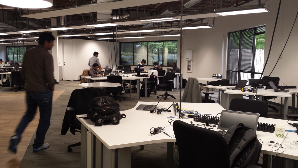 An office space with people working at white desks equipped with laptops and cables, featuring ceiling lights and large windows, creating a collaborative environment.