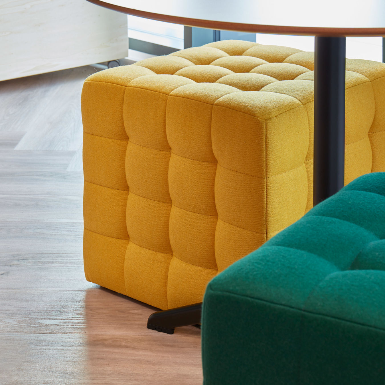 yellow cube-shaped ottoman with a tufted texture sits beside a green ottoman, under a circular table, on a light wooden floor, creating a modern, cozy feel