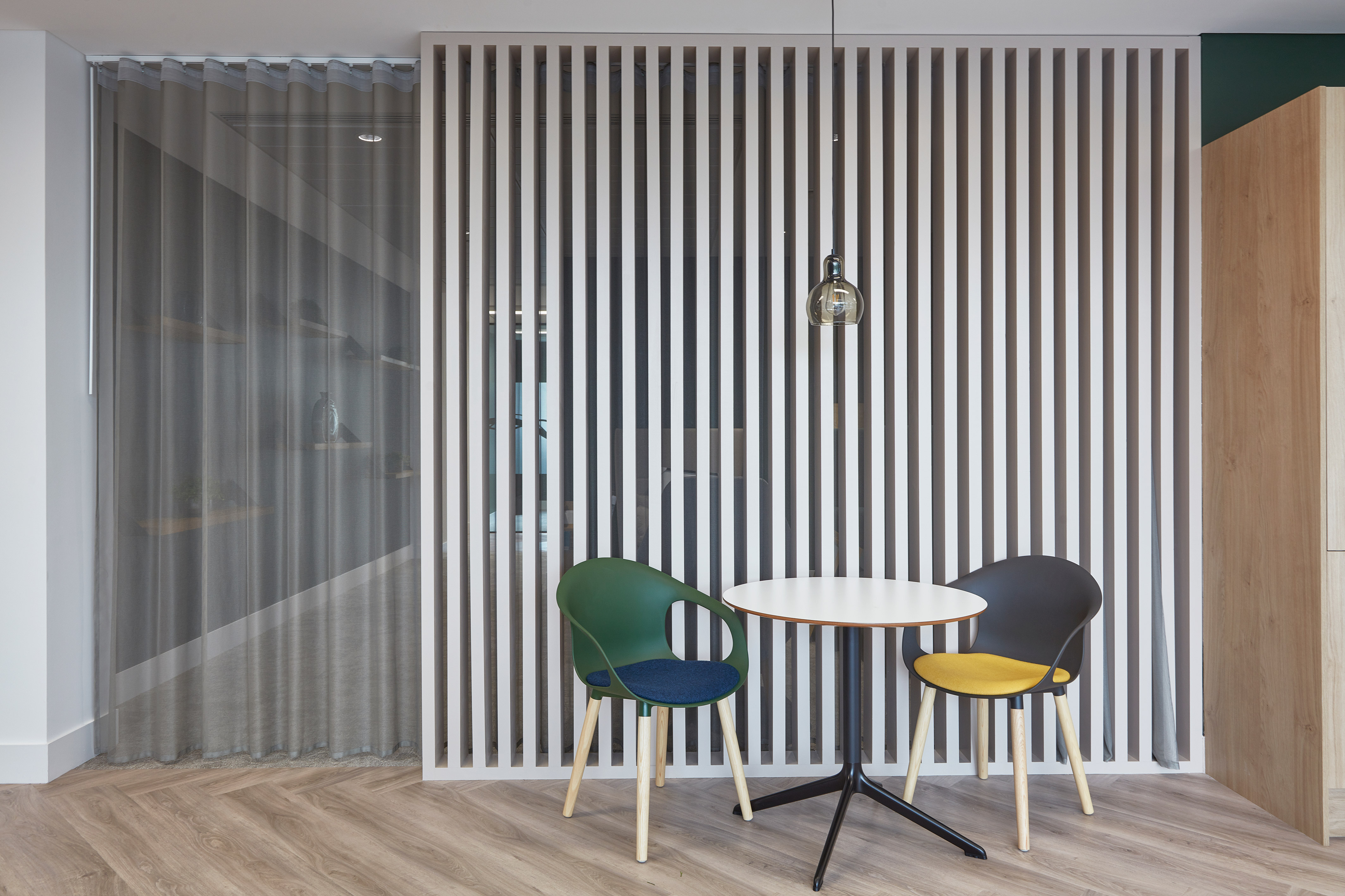 Two colorful chairs face a small round table under a single pendant light, set against a vertical slat partition in a modern, minimalist room.