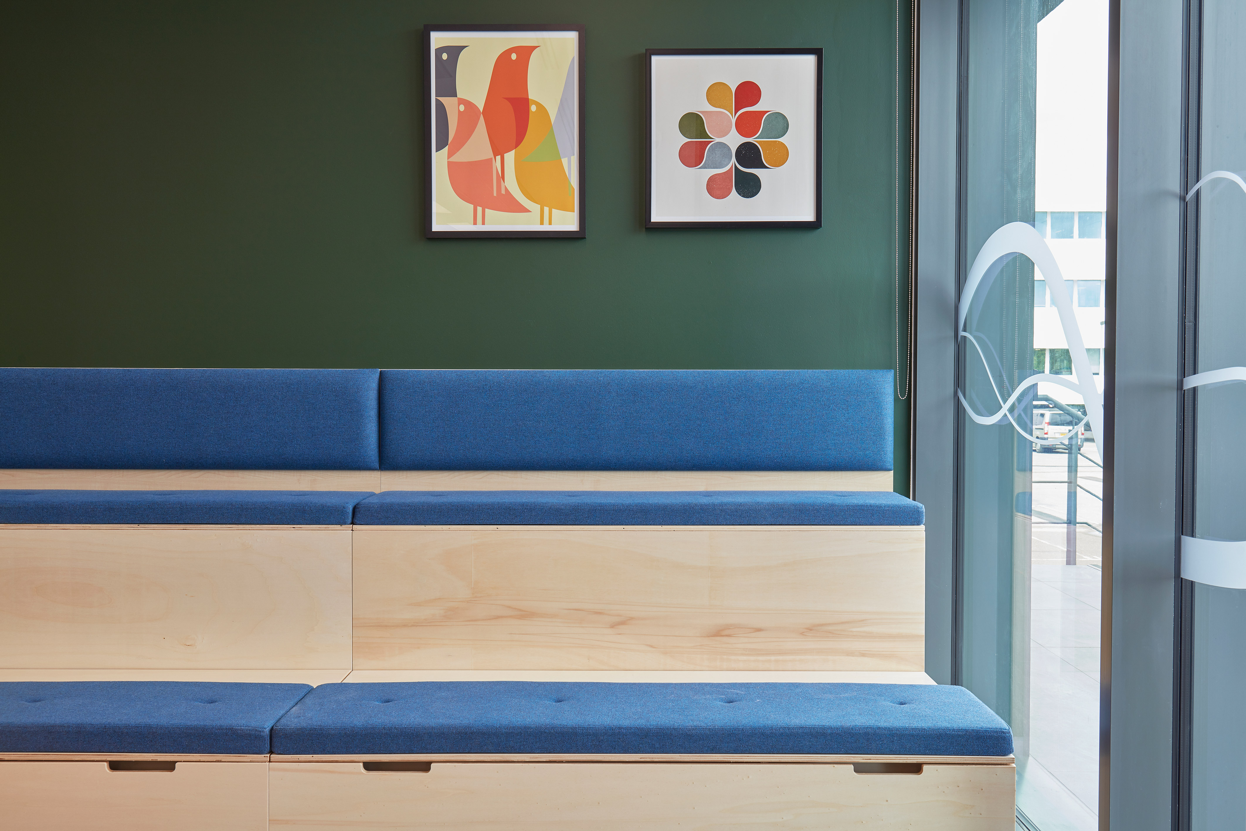 A modern lobby features wooden tiered seating with blue cushions against a dark green wall adorned with abstract art. Large windows add natural light to the space.