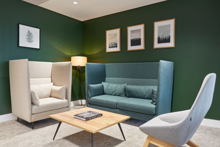 Modern lounge with green walls features a beige armchair, teal sofa, and grey chair. A wooden table holds a book. Warm lighting and framed art create a cozy ambiance