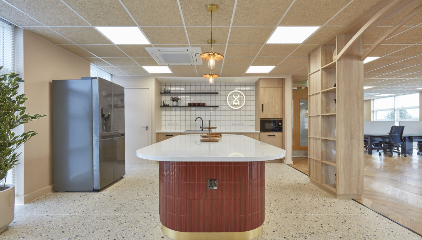 Kitchen island with a white countertop and red base stands in an office kitchen area, featuring a fridge, wooden cabinets, a neon clock on tiled wall, and pendant lights.