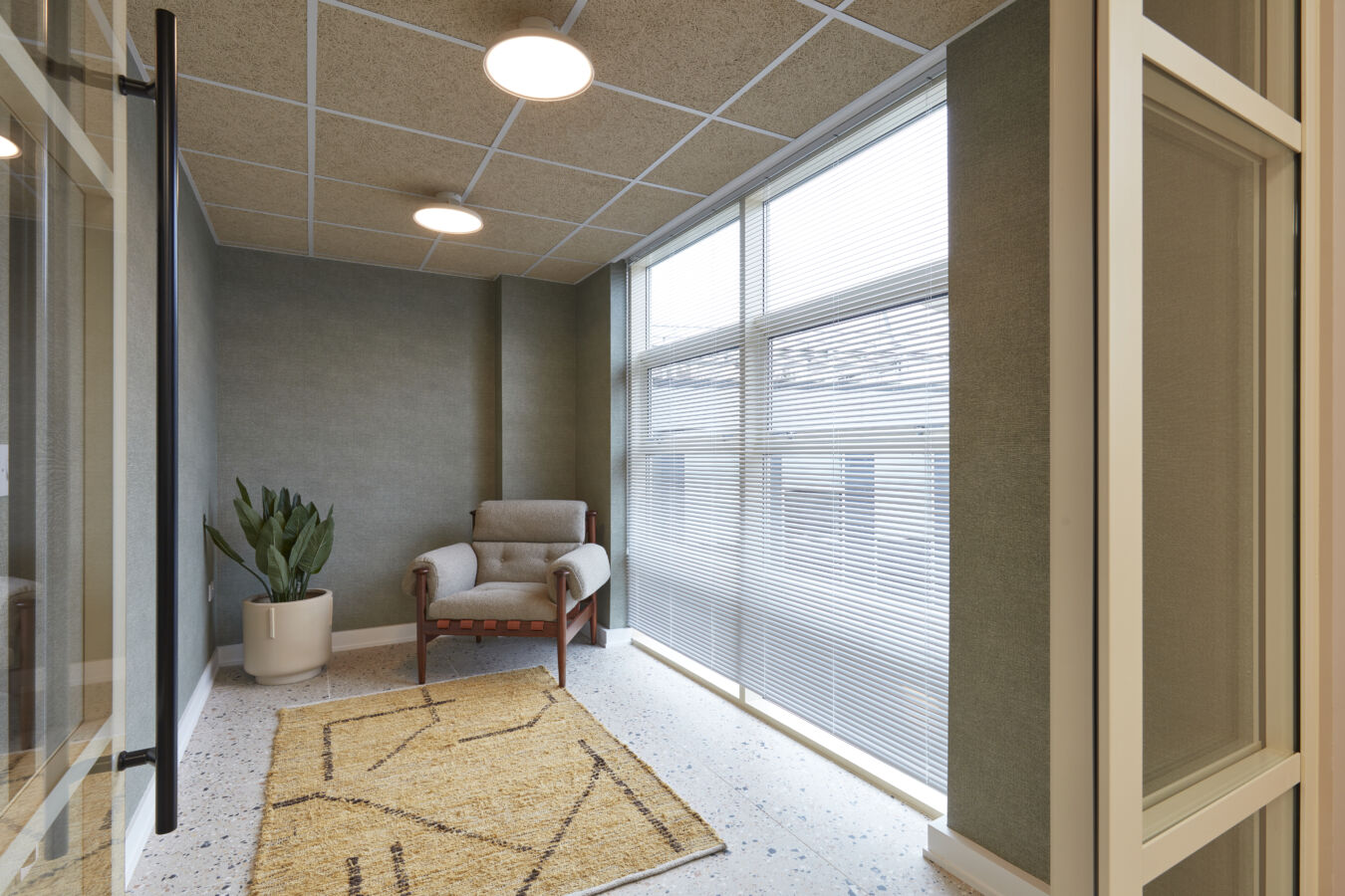 Quiet corner with armchair, patterned rug, large windows with blinds, and potted cactus in a softly lit space