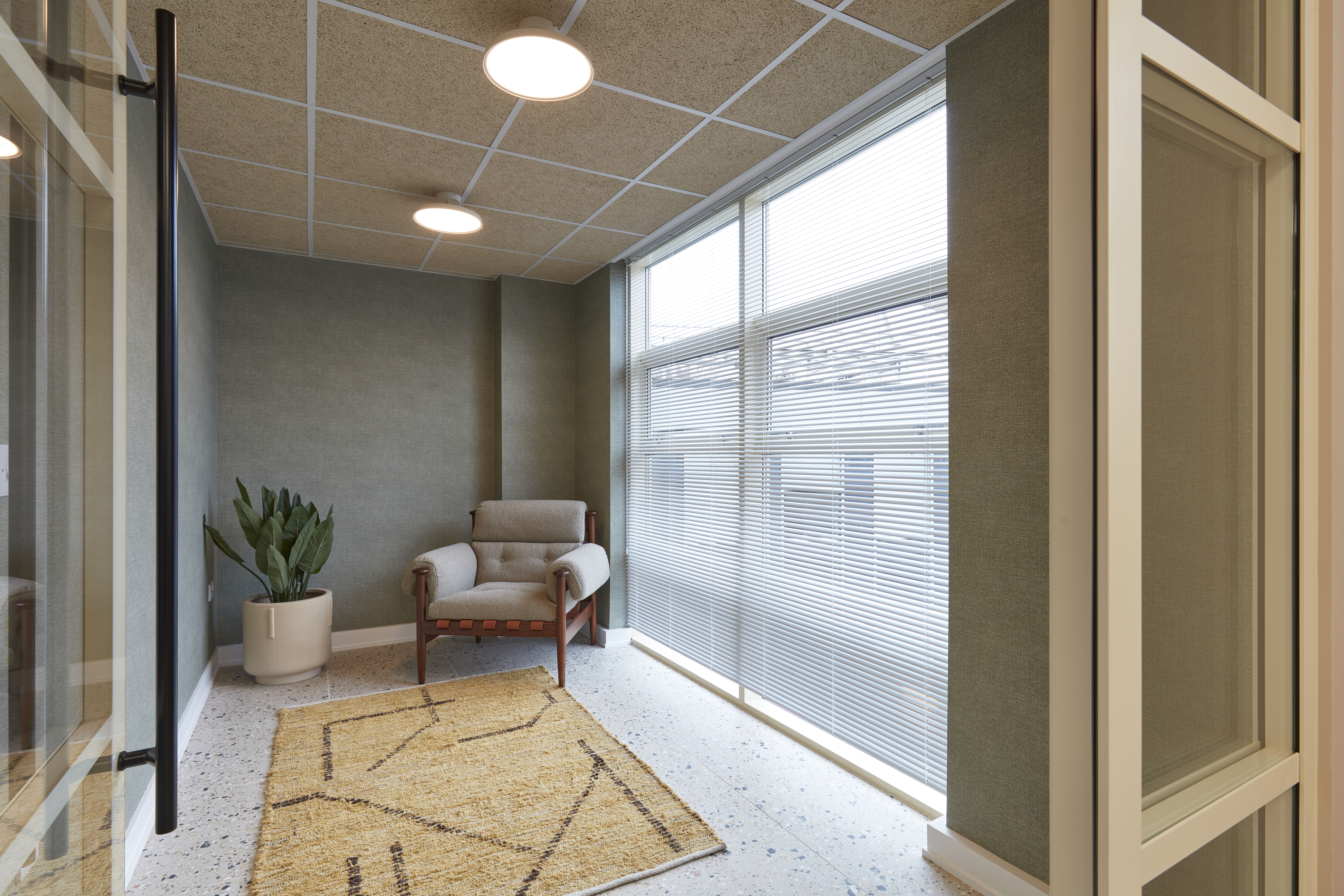 Quiet corner with armchair, patterned rug, large windows with blinds, and potted cactus in a softly lit space