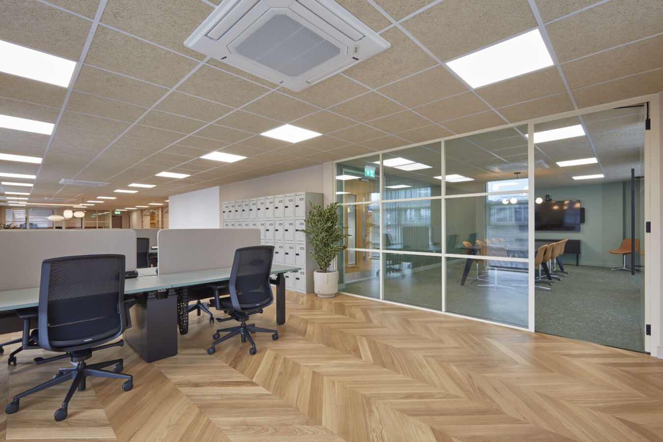 Office space featuring ergonomic chairs and desks with privacy panels, surrounded by light wood flooring, glass-walled meeting room, overhead fluorescent lighting, and white lockers on the side.