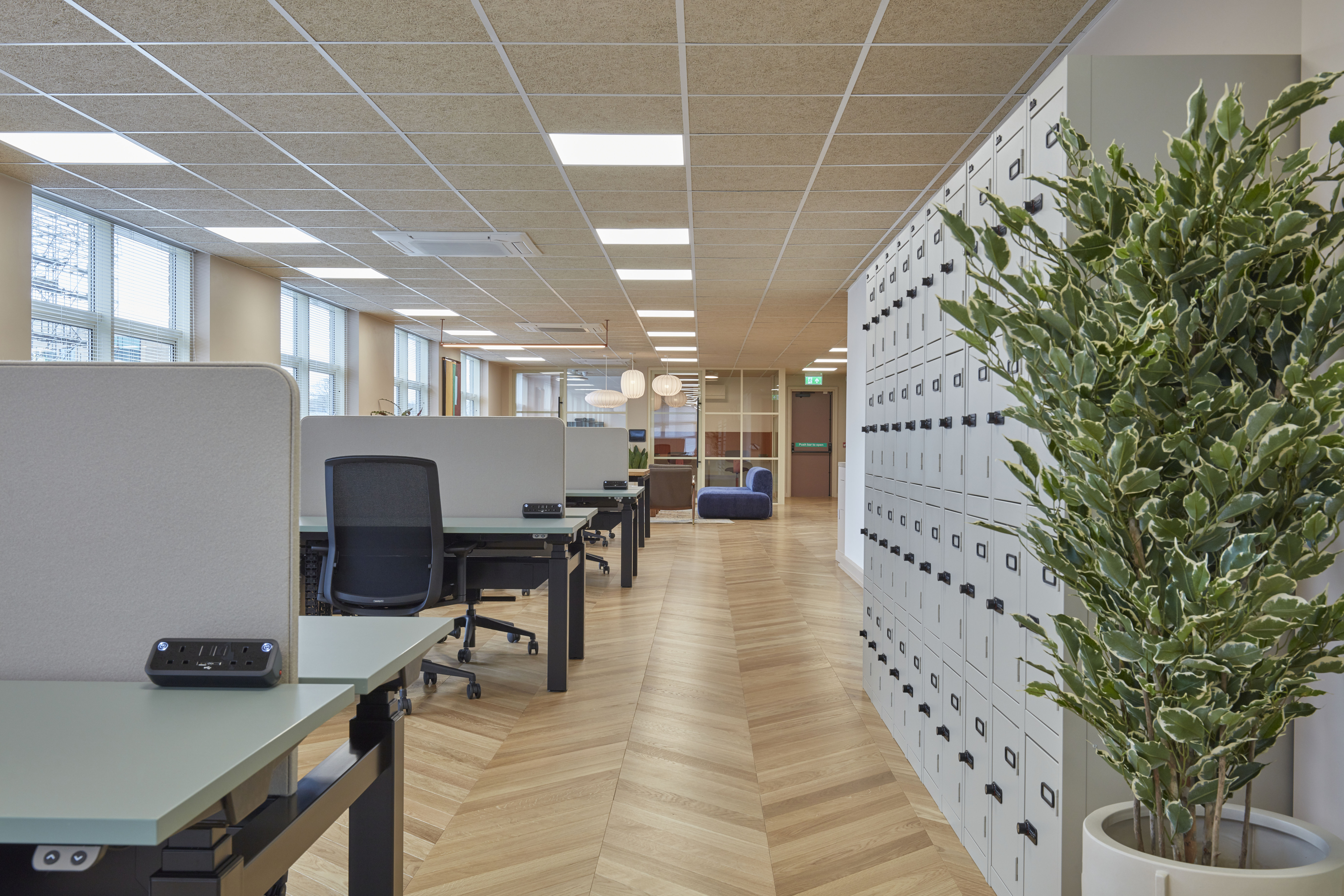 Open office space featuring workstations, black mesh chairs, wooden flooring, and rows of white storage lockers.