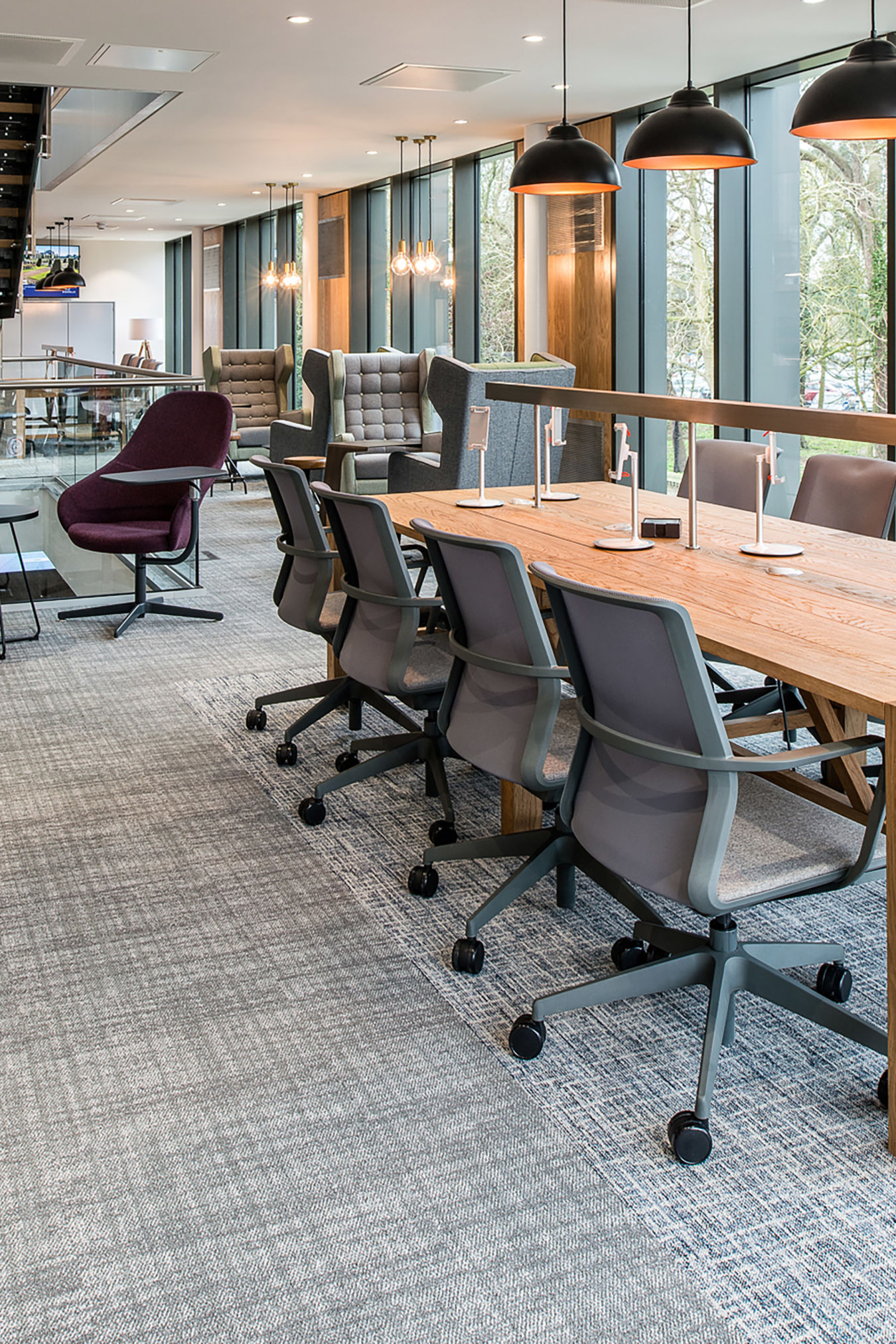Modern office space with a long wooden table, gray swivel chairs, and hanging black lamps. Large windows let in natural light, creating an airy atmosphere