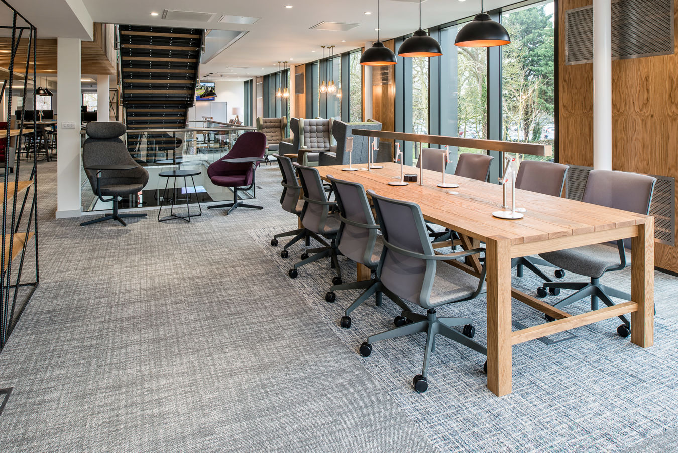Modern office space with a long wooden table, gray swivel chairs, and hanging black lamps. Large windows let in natural light, creating an airy atmosphere