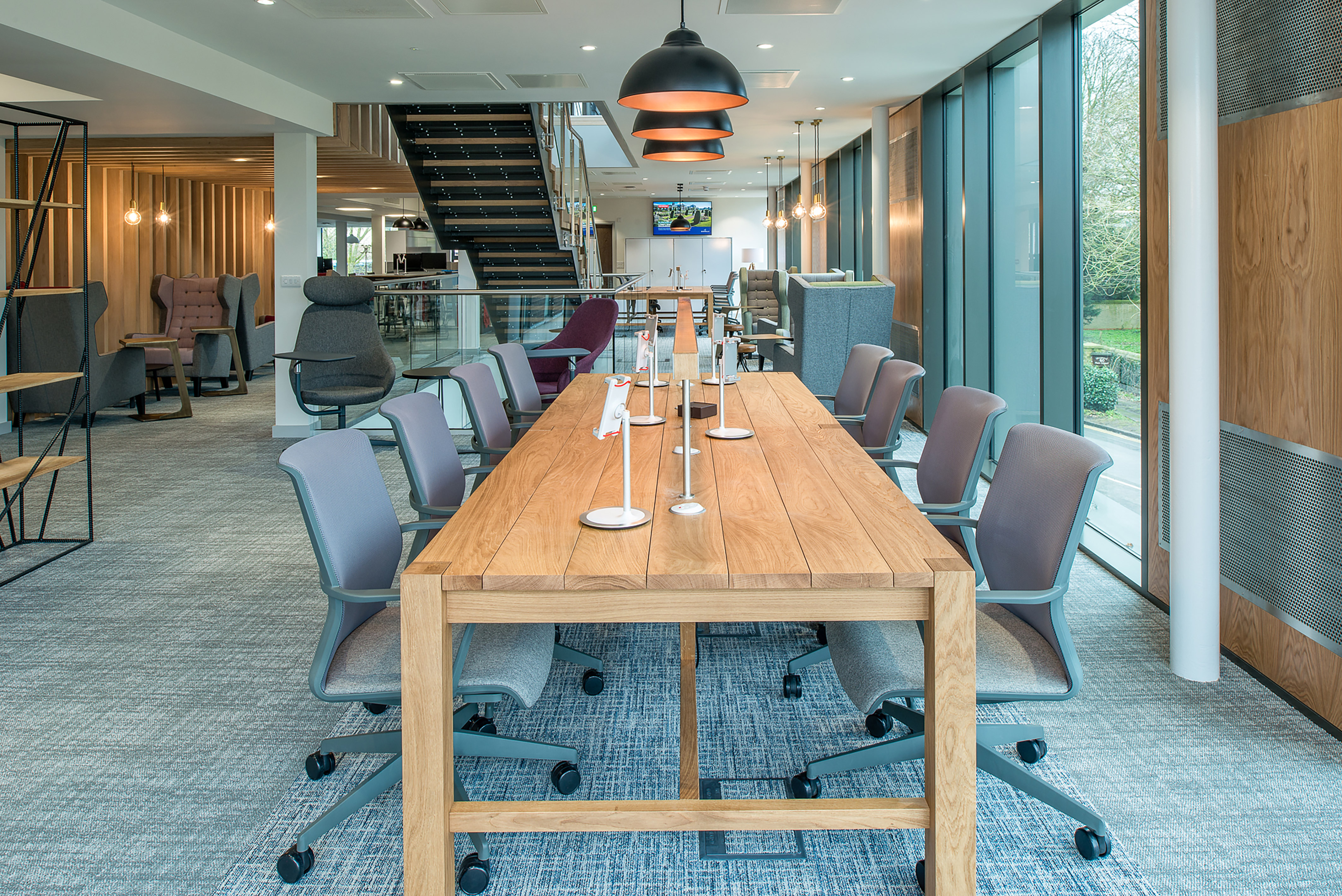 Modern office space with wooden tables and ergonomic chairs arranged in a row. Pendant lights hang overhead, and large windows provide natural light. A staircase and casual seating are in the background.