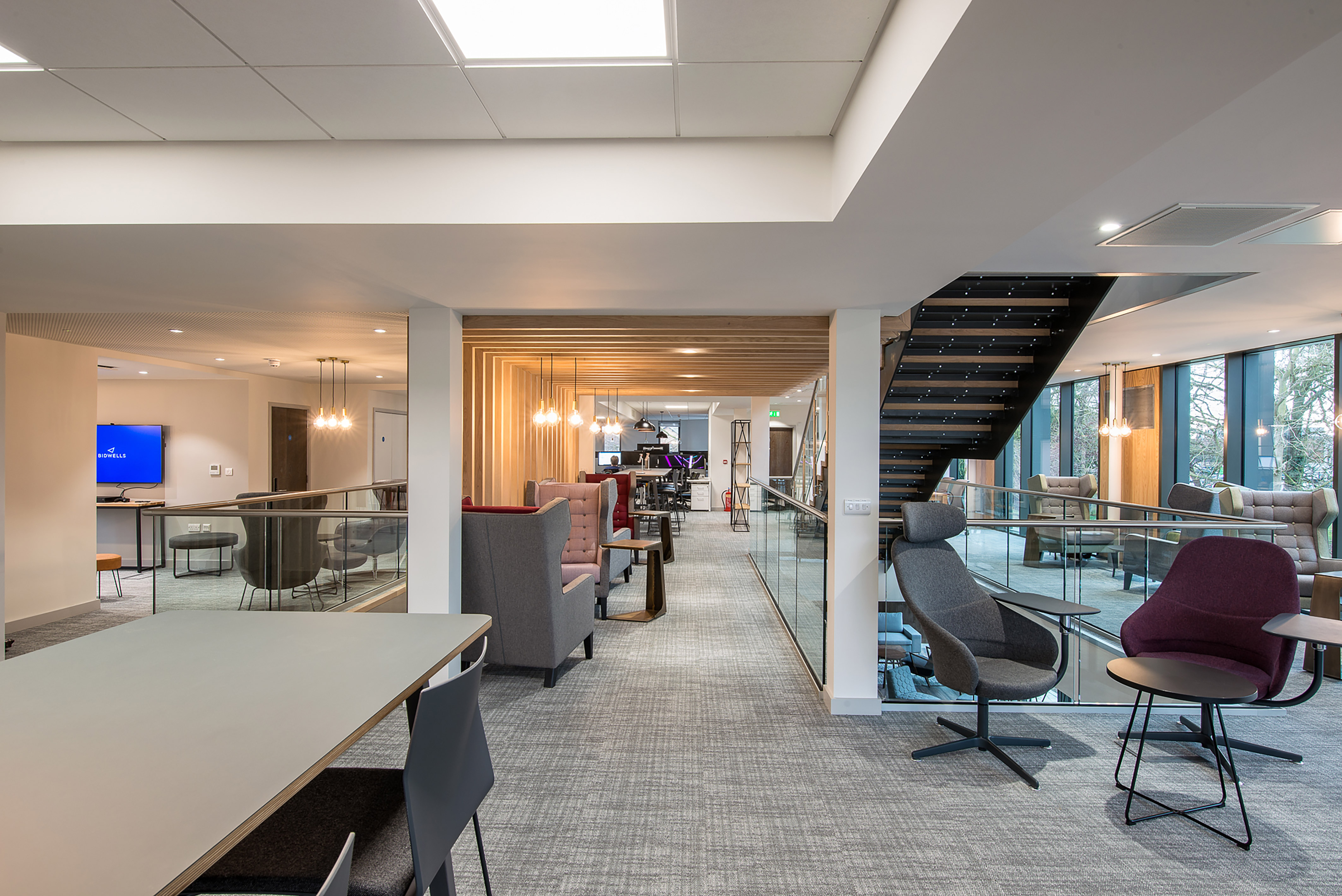 Modern office space with stylish chairs and tables, glass partitions, and a staircase, featuring a wall-mounted screen displaying "VIDEOLLES" in a well-lit, open-floor design.