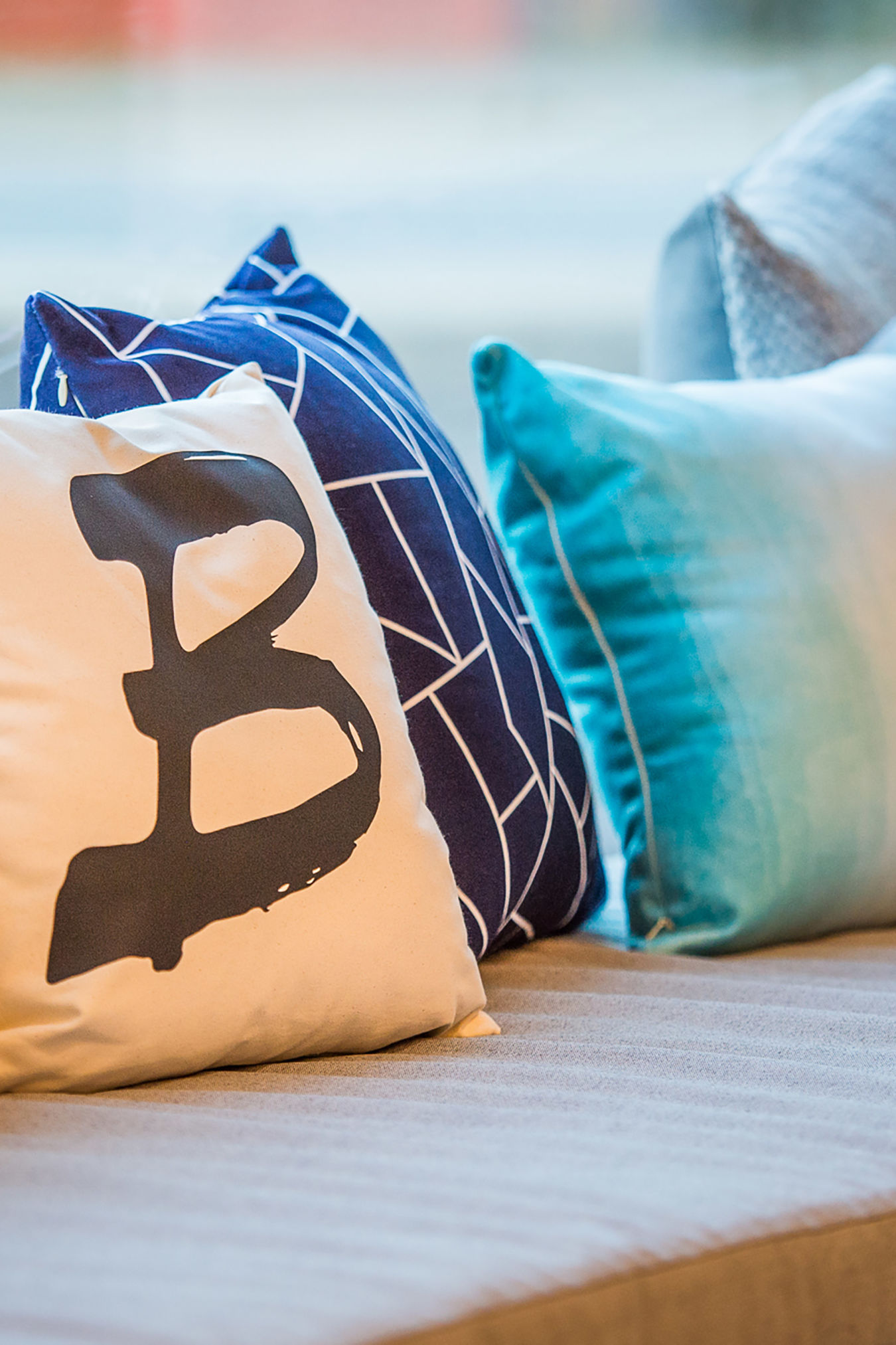 Cushions rest on a sofa in a brightly lit room; one cushion features a black letter "B" on a white background, surrounded by patterned blue and gray pillows.