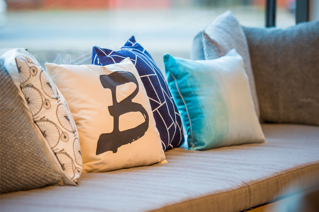 Cushions rest on a sofa in a brightly lit room; one cushion features a black letter "B" on a white background, surrounded by patterned blue and gray pillows.