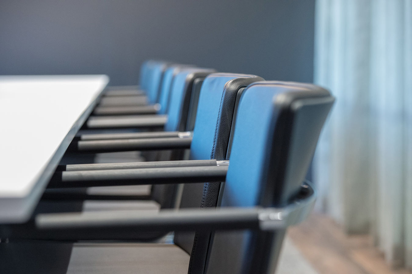 Conference chairs with blue upholstery are neatly aligned along a sleek white table in a modern meeting room with grey walls and softly lit space.