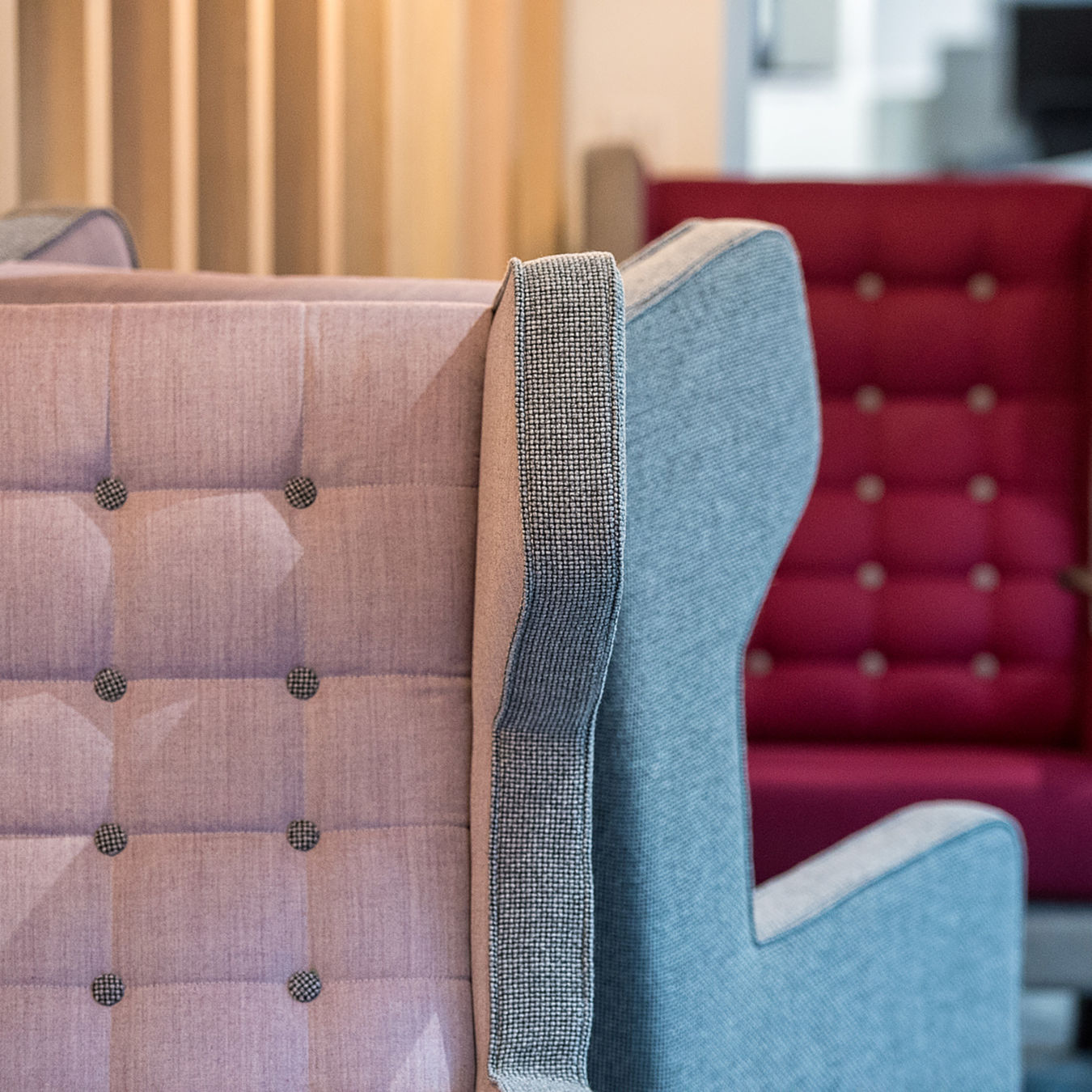 Close-up of two upholstered armchairs in an interior setting. The foreground chair is light pink with a tufted design; the background chair is red. Cozy and modern