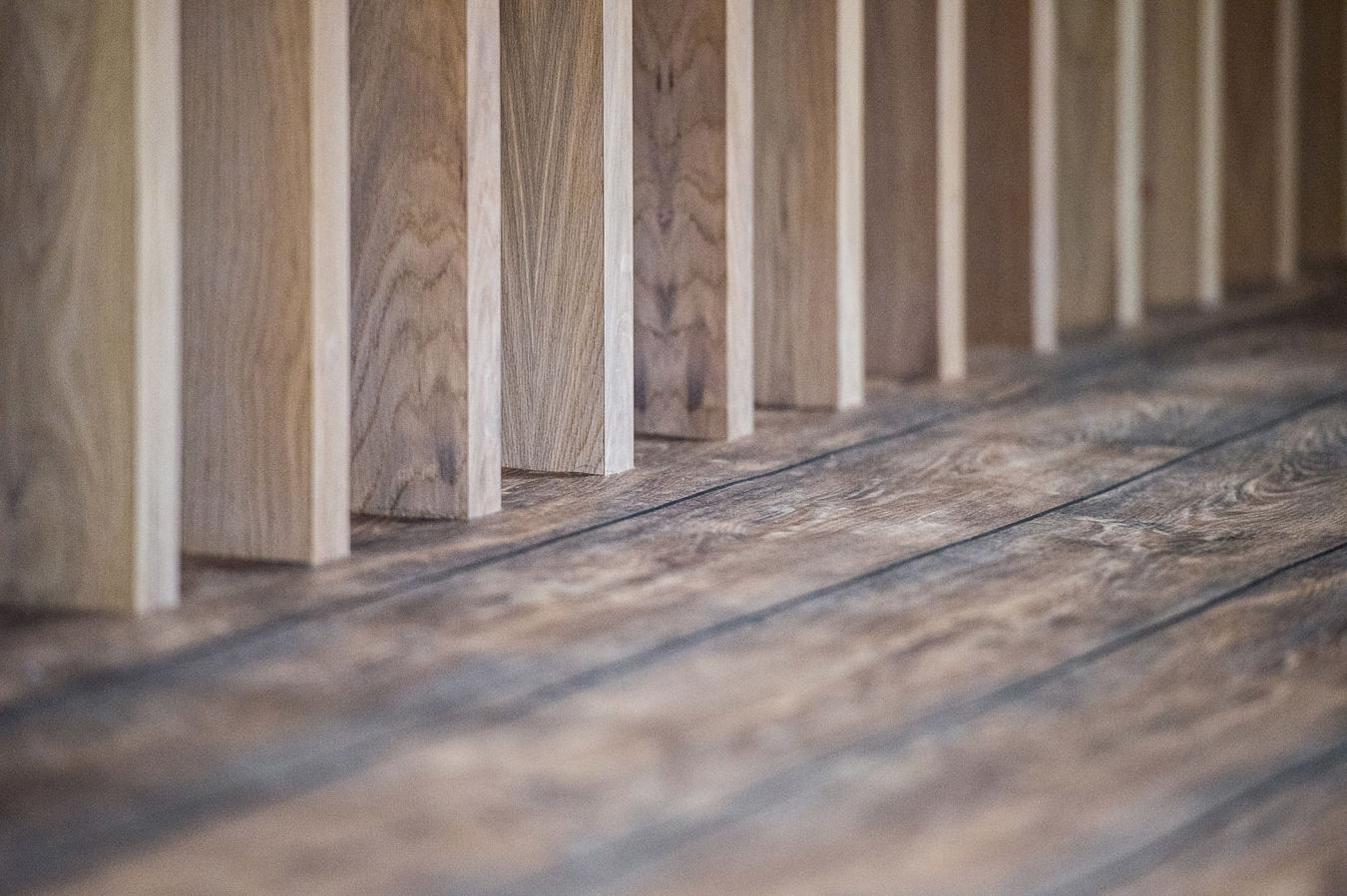 Wooden planks positioned vertically form a partition, casting shadows on a textured wooden floor, creating a pattern in a softly lit interior.