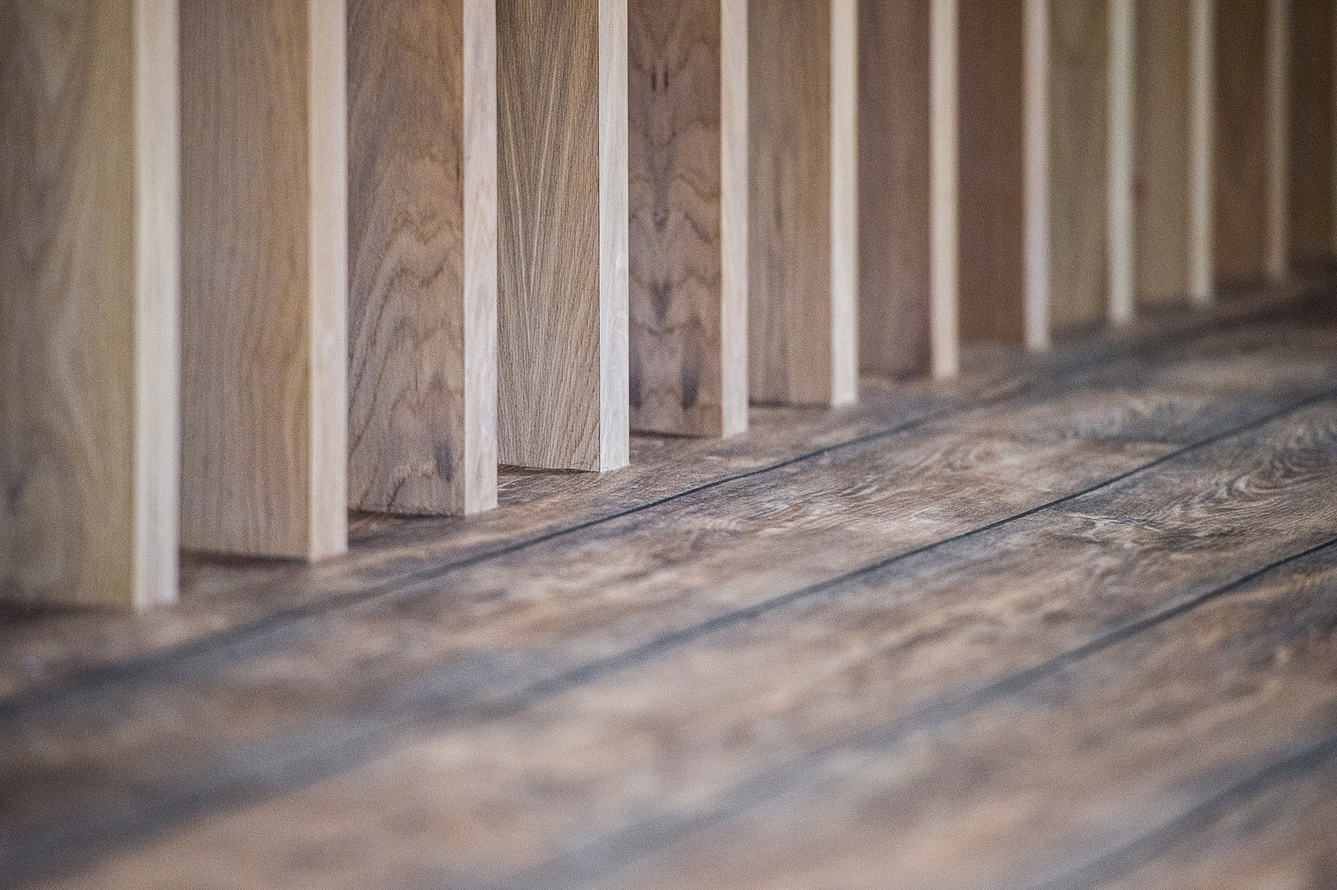 Wooden planks positioned vertically form a partition, casting shadows on a textured wooden floor, creating a pattern in a softly lit interior.