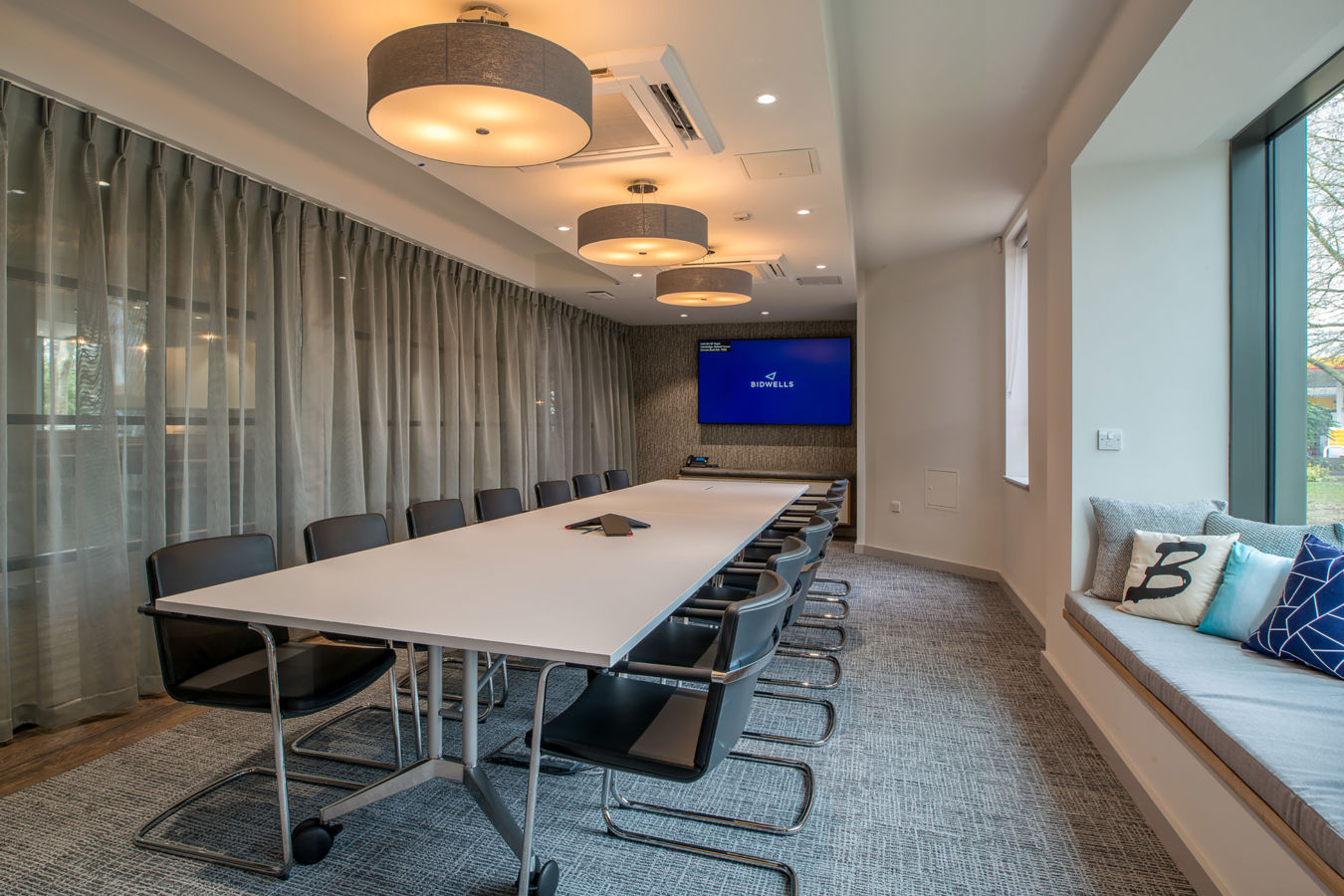 Modern conference room with a long white table, black chairs, and large screen on the wall. Soft lighting and window seat with decorative pillows