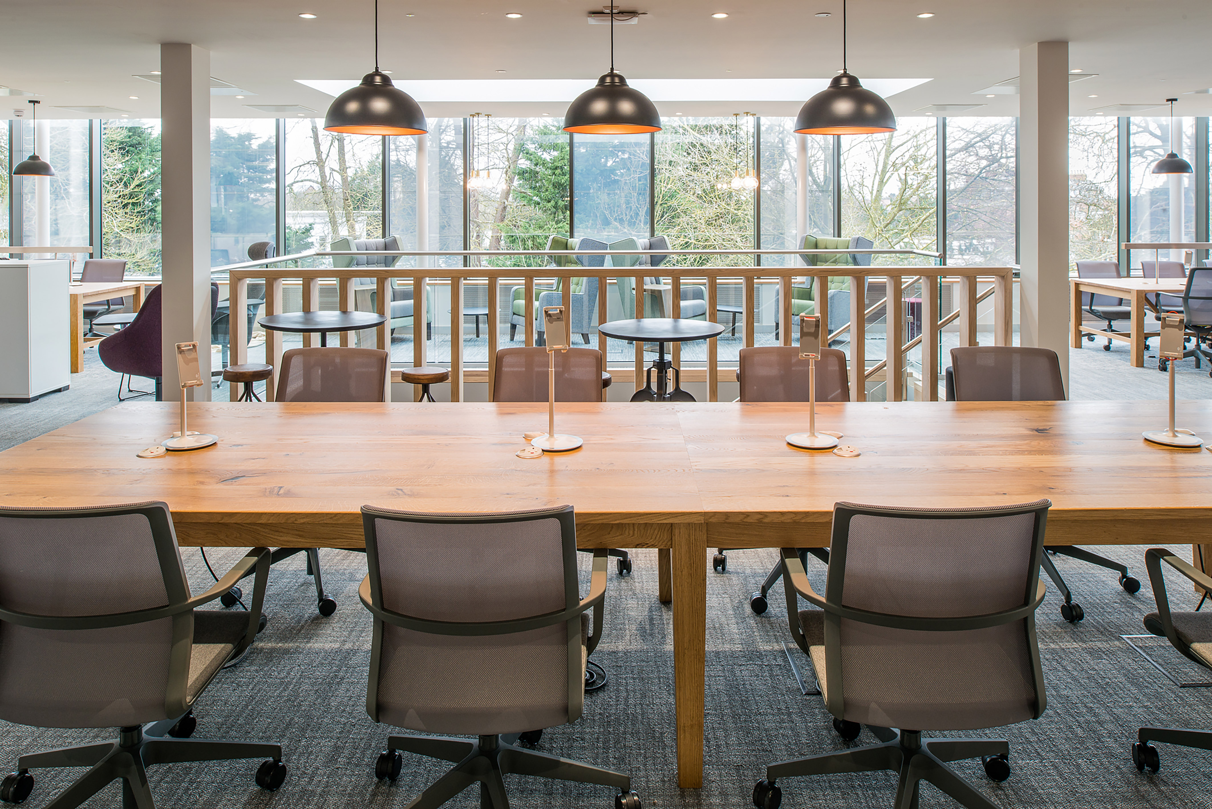 Modern office space with a large wooden table, gray chairs, and pendant lights. Floor-to-ceiling windows reveal trees outside, creating a serene atmosphere