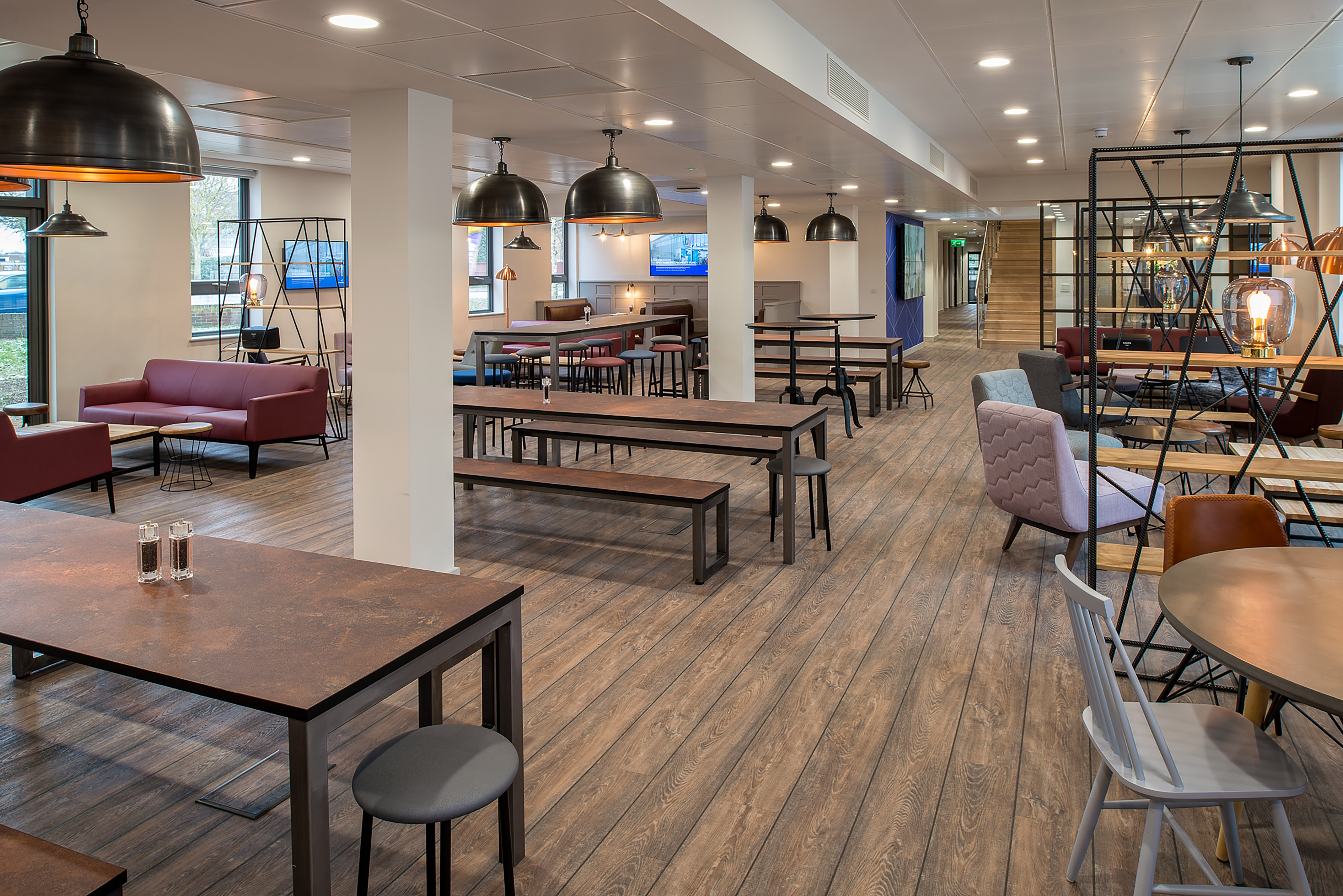 A modern office lounge features wooden tables and benches, with hanging industrial lights. Seating includes sofas and chairs, and shelves hold decorative items in a well-lit, spacious area.