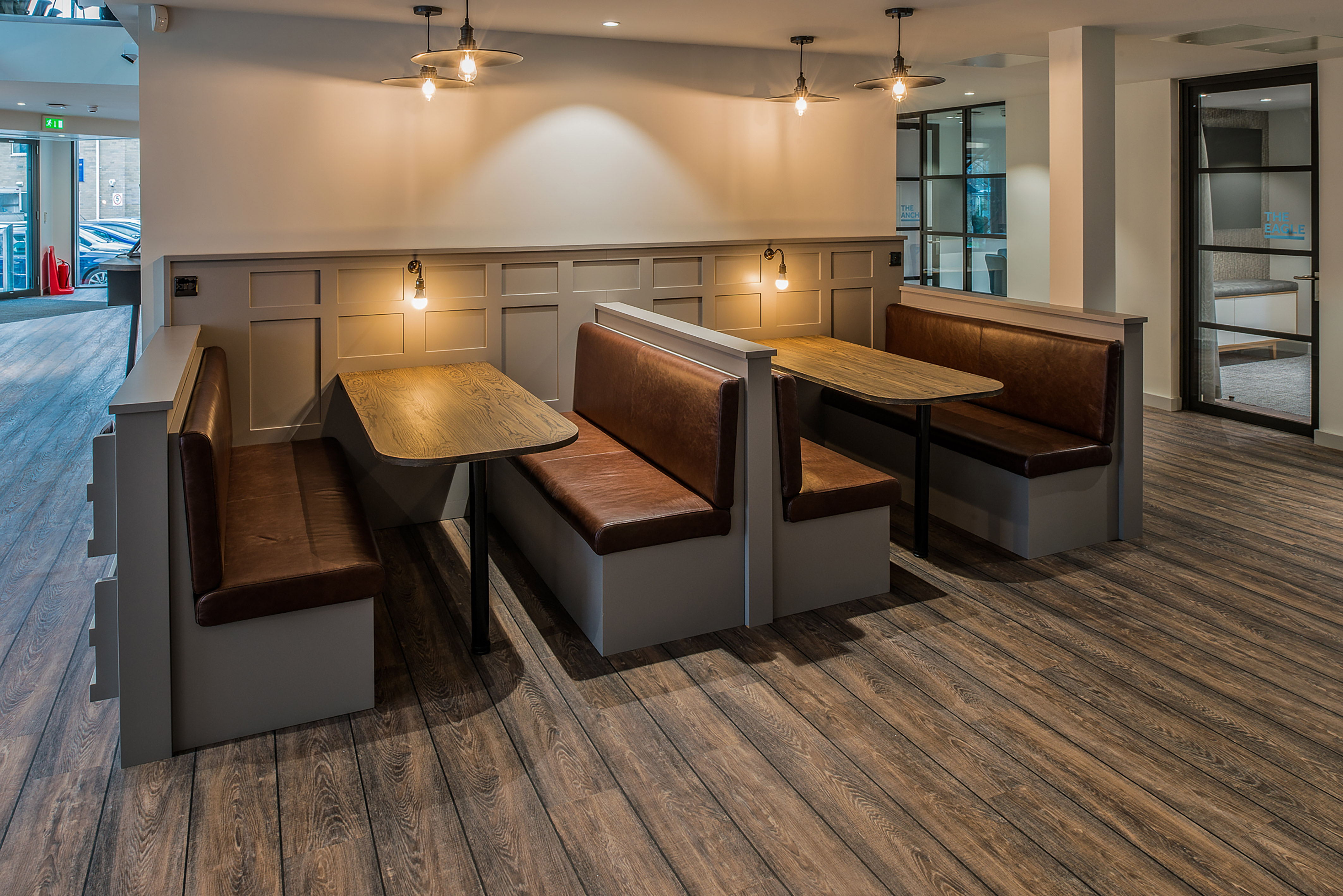 Modern cafe interior with wooden flooring, featuring two booth seating areas with brown leather benches and wooden tables. Warm pendant lights enhance the cozy atmosphere