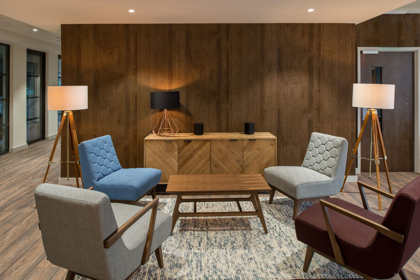 Stylish modern lounge with four mid-century armchairs around a wooden coffee table. Warm lighting from tall lamps, wood panel walls, and cozy ambiance
