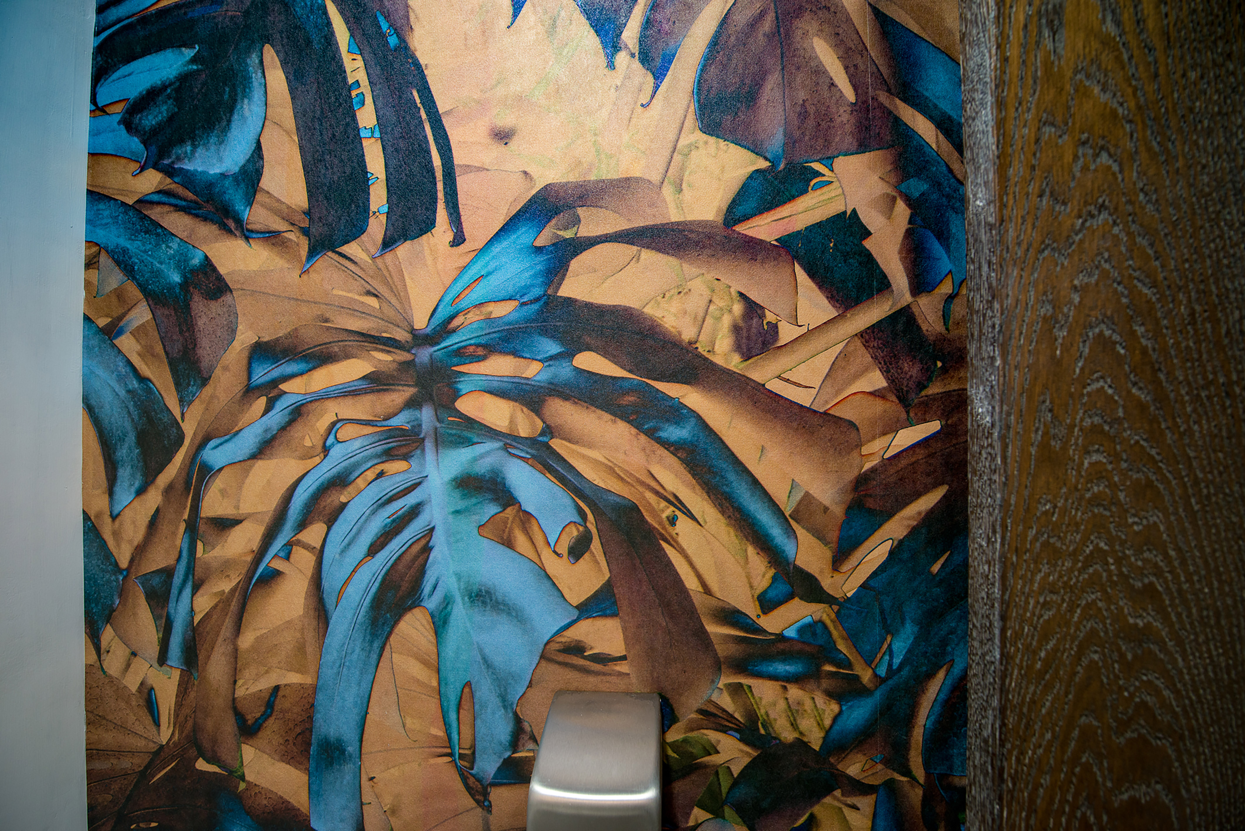 Colorful tropical leaf mural decorates an indoor wall, with large blue and brown leaves overlapping. A metal door handle is partially visible on the right.