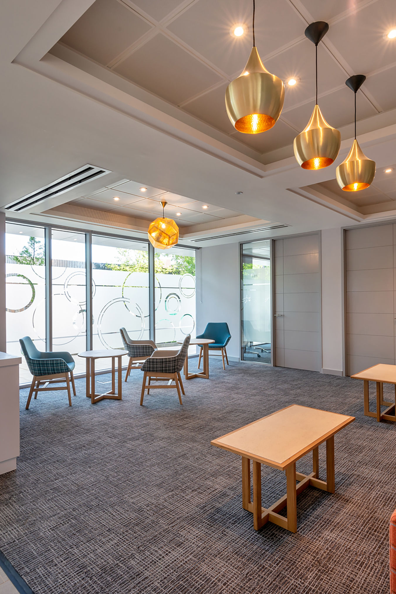 Modern lobby features wooden chairs and tables under warm pendant lights, with large windows displaying frosted circular patterns and an office door adjacent.