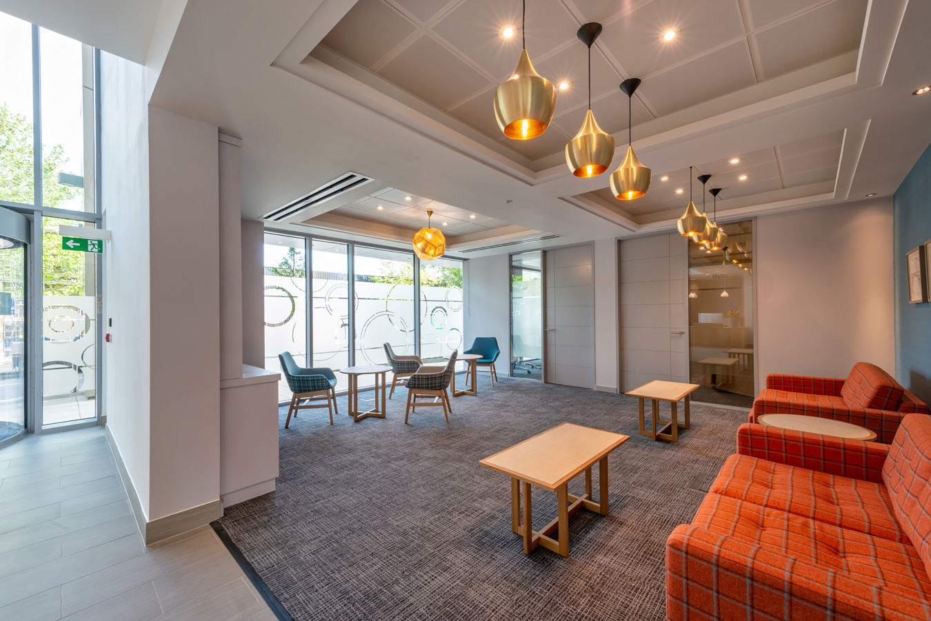 Modern lobby features wooden chairs and tables under warm pendant lights, with large windows displaying frosted circular patterns and an office door adjacent.