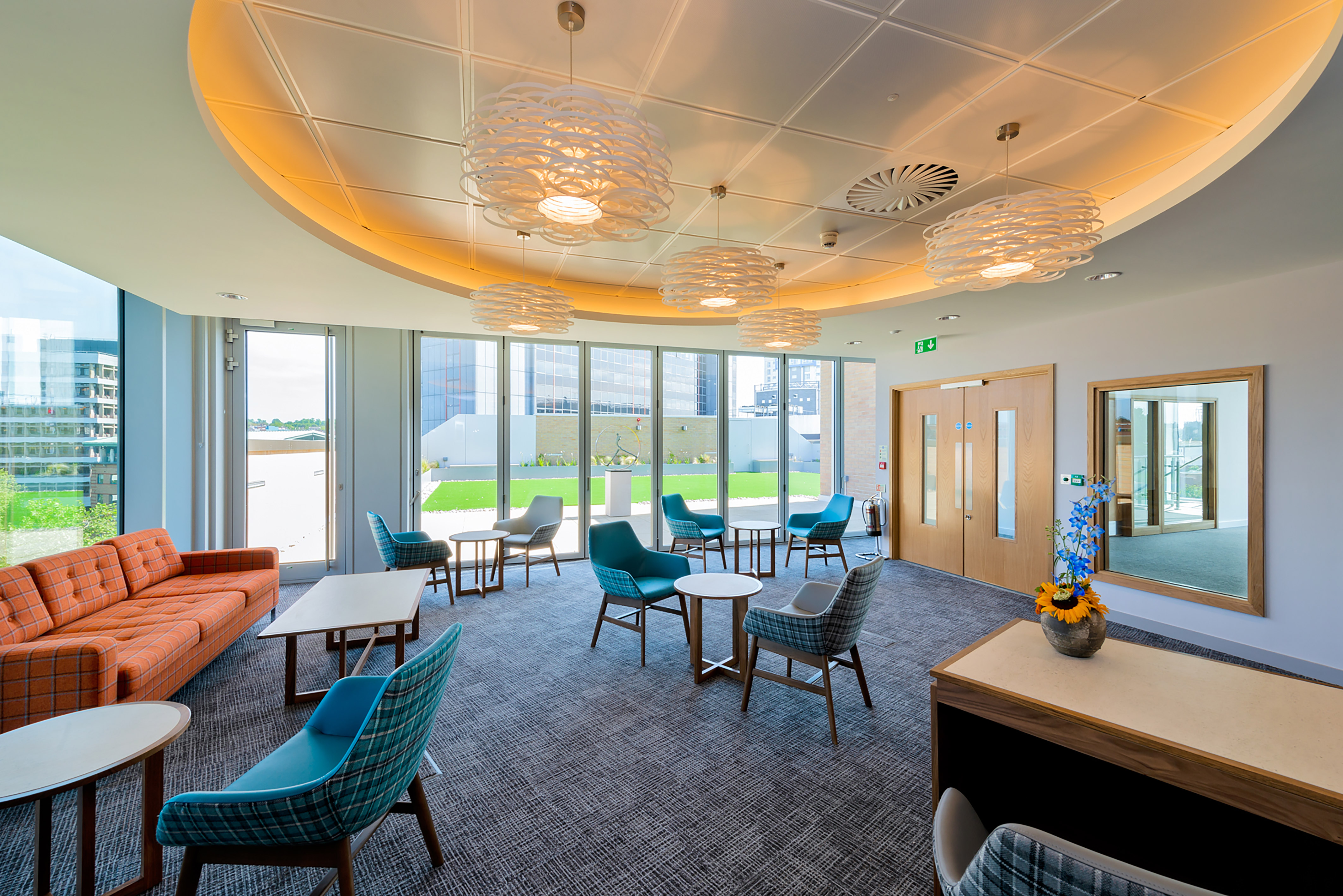 A modern lounge showcases colorful chairs and tables arranged around an orange sofa, under artistic ceiling lights, with floor-to-ceiling windows revealing an outdoor view.