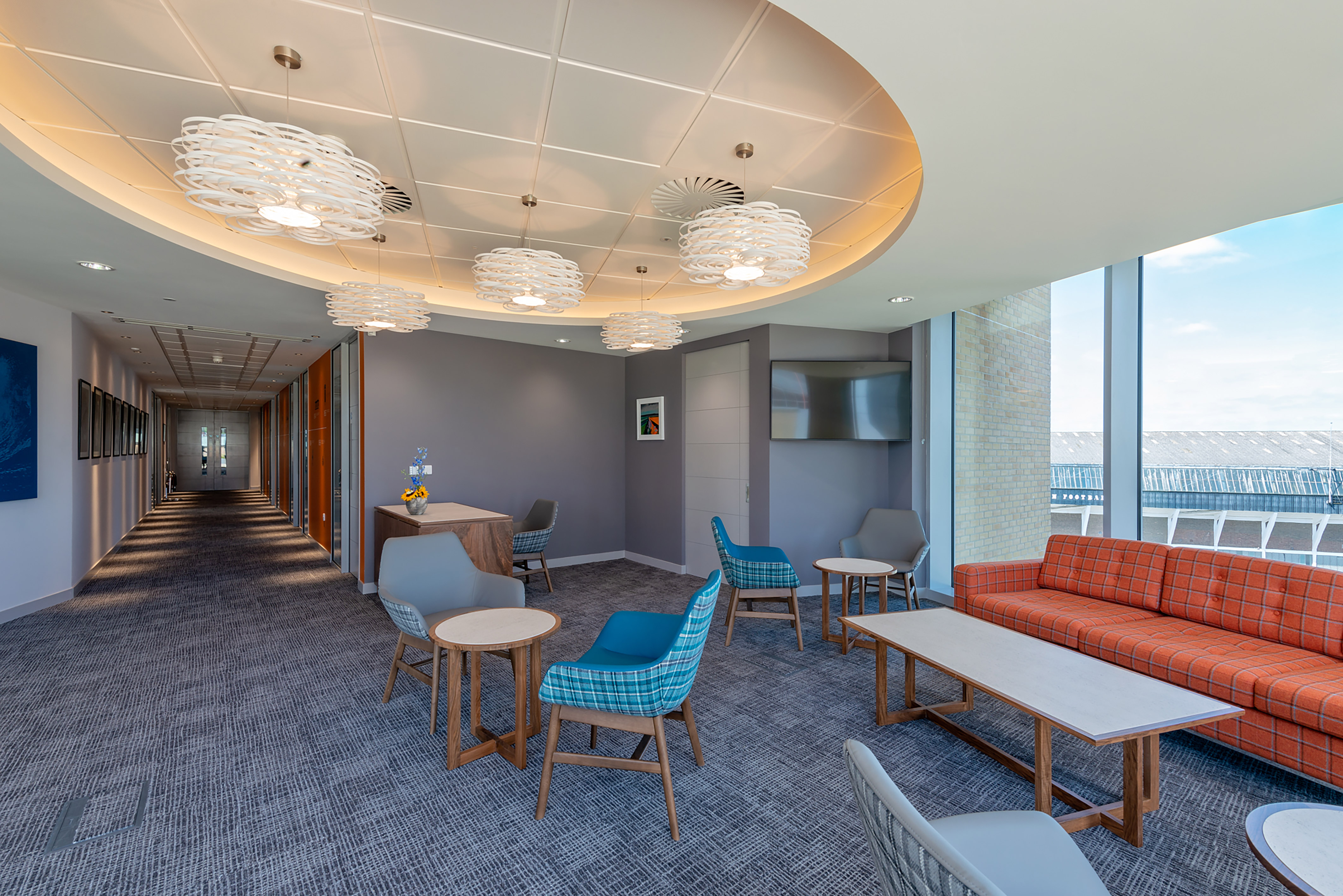 Lobby area with circular lighting, chairs, tables, and an orange plaid sofa; adjacent to a long corridor with artwork, set in an office environment with large windows revealing an outdoor view.